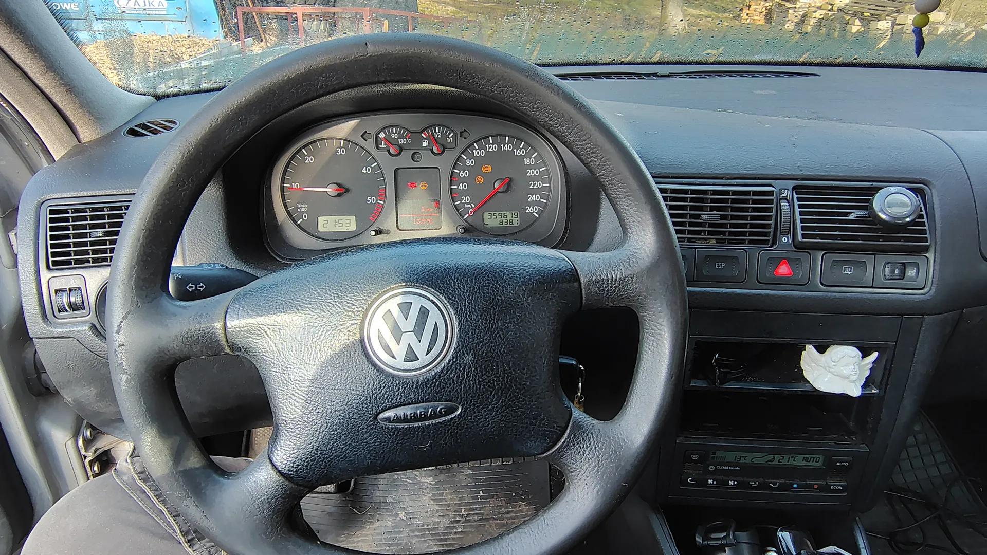 Wnętrze samochodu marki Volkswagen z widocznym kierownicą z logo VW i napisem AIRBAG. Tablica przyrządów zawiera wskaźniki prędkości (do 260 km/h), obrotomierz, cyfrowe wyświetlacze z wartościami 2153 i 359679.838 oraz ikony ostrzegawcze. Na desce rozdzielczej widoczne są przyciski ESP, czerwony przycisk alarmu, wentylacyjne otwory i radio z wyświetlaczem. Na środku deski rozdzielczej umieszczona jest biała figurka anioła. Przez przednią szybę widoczny zewnętrzny krajobraz z niebieską konstrukcją i ogrodzeniem. W tle widoczne są elementy kabiny, w tym kluczyki w stacyjce.