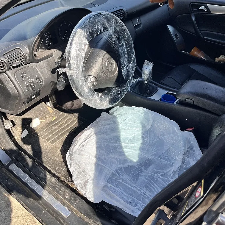 Wnętrze samochodu marki Mercedes-Benz z perspektywy otwartych drzwi kierowcy. Kierownica z logo marki jest owinięta przezroczystą folią ochronną. Na siedzeniu kierowcy znajduje się duża, biała plastikowa folia. Widoczna jest deska rozdzielcza z zegarami, lewarek zmiany biegów oraz czarne elementy wykończenia wnętrza. Na progu samochodu widoczny jest napis 'Mercedes-Benz'.