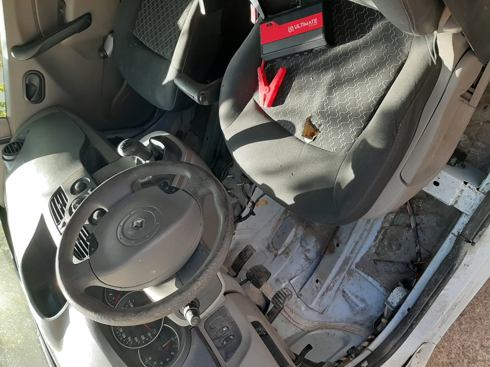 Wnętrze samochodu z widoczną kierownicą marki Renault, deską rozdzielczą z licznikami, siedzeniem pasażera z uszkodzeniem (dziurą) oraz czerwonym klipsem i czarno-czerwonym pudełkiem z napisem ULTIMATE. Widać również otwarte drzwi, częściowo rozebrany panel podłogowy z odsłoniętymi przewodami oraz fragment zewnętrznej powierzchni po prawej stronie.