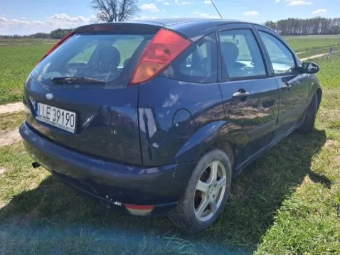 Ciemnoniebieski samochód osobowy typu hatchback (Ford Focus) widziany od tyłu z boku, zaparkowany na trawie. Widoczna tablica rejestracyjna z numerem LE 39190. Tło stanowi pole, drzewa i niebo z chmurami. Odbicia na karoserii sugerują otoczenie z drzewami i niebem.
