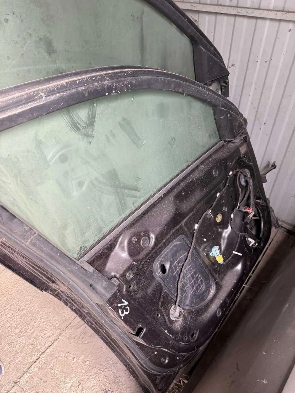 Zdjęcie przedstawia uszkodzoną część pojazdu, prawdopodobnie drzwi, z zielonkawym szybą. Czarna obudowa posiada widoczne uszkodzenia, odsłonięte przewody elektryczne oraz napis "13" na dolnej części. W tle metalowa, pionowo zarysowana ściana z blachy falistej i betonowa podłoga. Całość pokryta jest kurzem i śladami zarysowań.