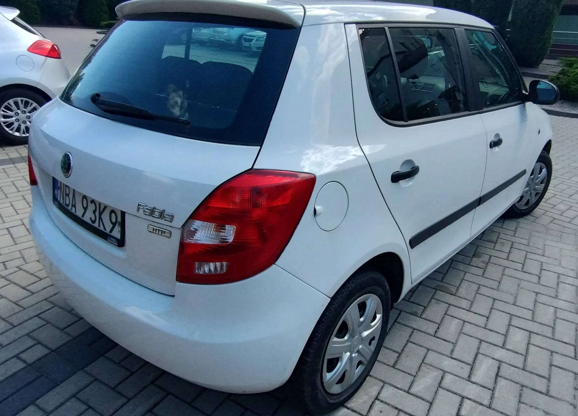 Biały samochód marki Skoda Fabia HTP zaparkowany na kostce brukowej. Widoczny jest tył i prawy bok pojazdu. Samochód ma tablicę rejestracyjną o numerze NBA 93K9 oraz oznaczenia modelu 'Fabia' i 'HTP' na klapie bagażnika.