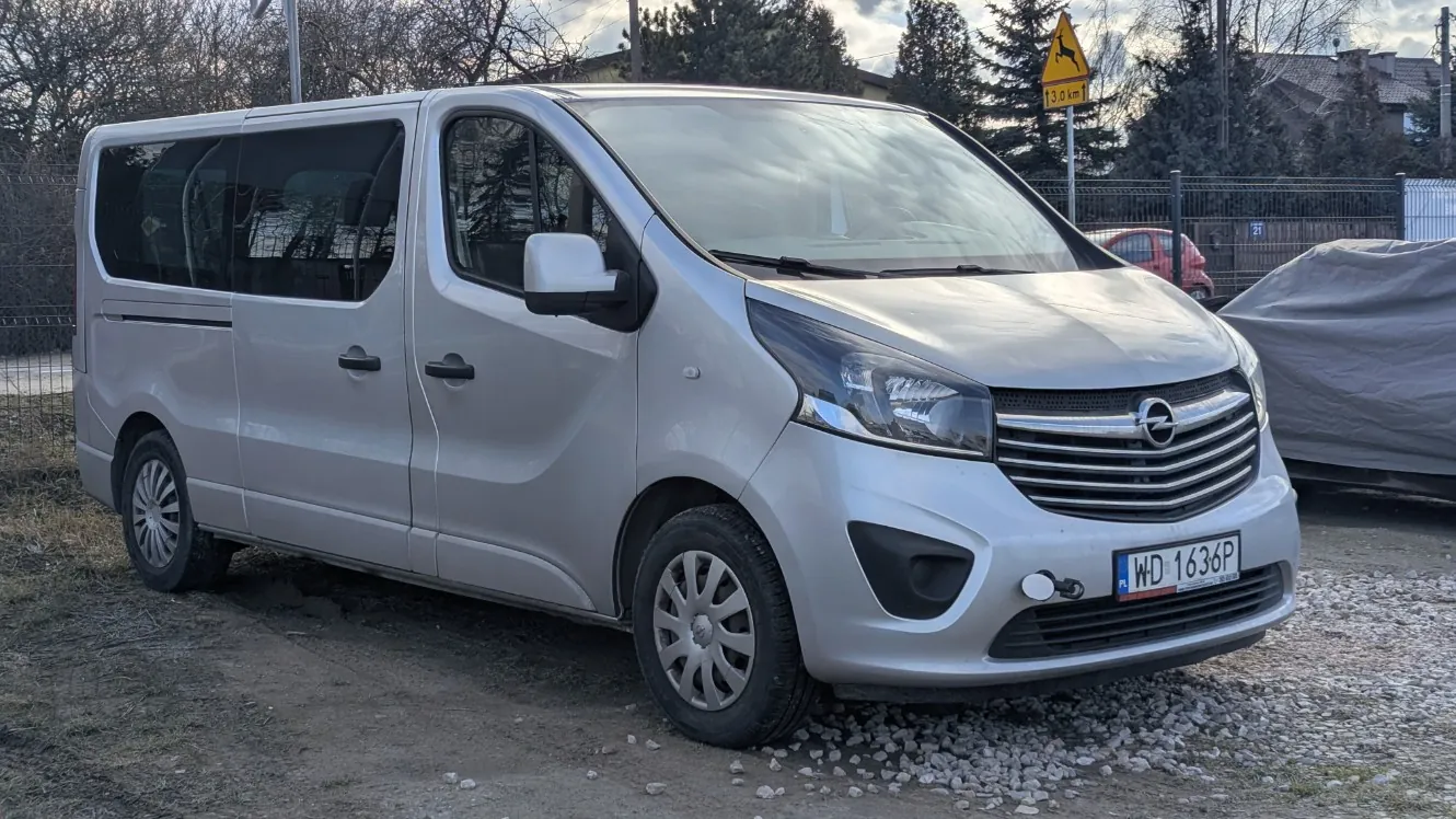 Srebrny samochód dostawczy marki Opel z tablicą rejestracyjną WD 1636P, zaparkowany na podwórku z żwirem i ziemią. W tle widoczny żółty znak drogowy z sylwetką jelenia i napisem 13.0 km, ogrodzenie, drzewa, czerwony samochód oraz zakryty pojazd. Niebo częściowo zachmurzone.