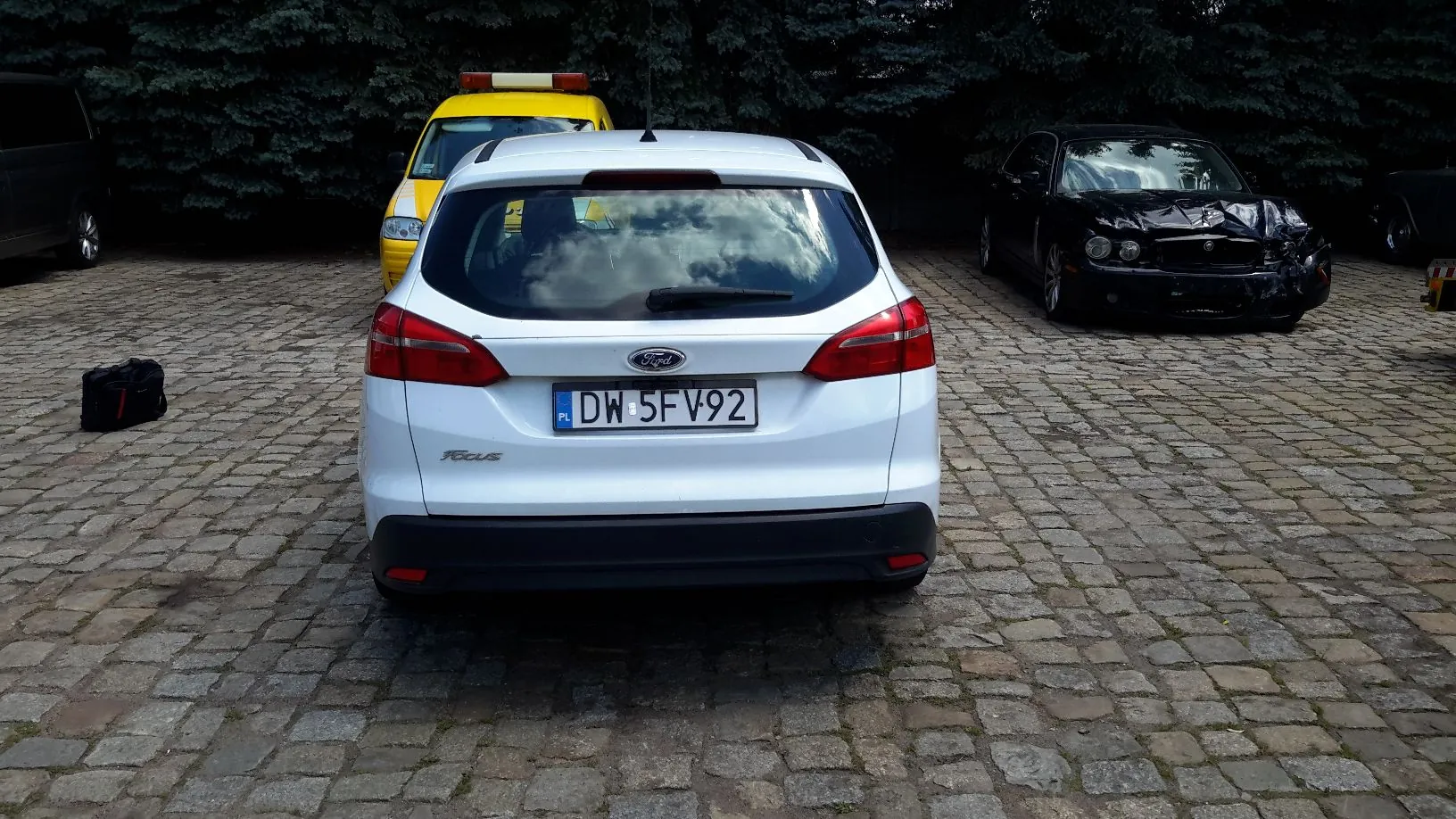 Widok z tyłu białego samochodu marki Ford Focus z polską tablicą rejestracyjną DW 5FV92, zaparkowanego na brukowanym placu. W tle po lewej stronie widoczny jest żółty samochód służbowy, a po prawej czarny samochód z poważnie uszkodzonym przodem. W tle znajduje się gęsty żywopłot z iglaków.