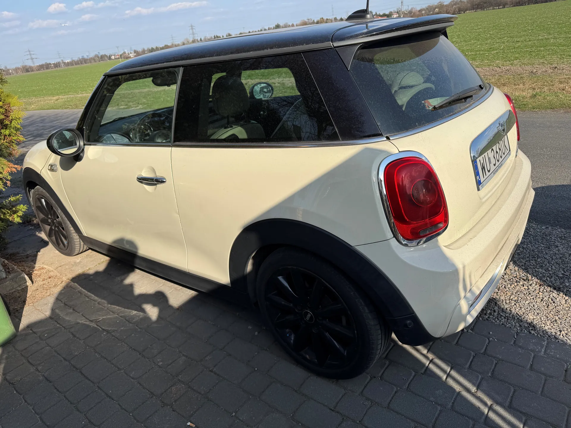 Biały samochód Mini Cooper z czarnym dachem zaparkowany na brukowanej powierzchni. Widok z tyłu i boku. Tablica rejestracyjna WU 368K. W tle zielone pole, niebo z chmurami oraz drzewa. Czarne felgi, czerwone światła tylne, lusterka boczne. Słoneczny dzień, cienie na ziemi.