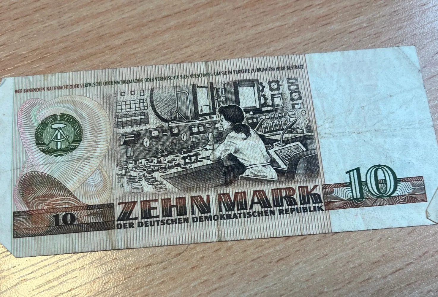 Banknot o nominale 10 marek z Niemieckiej Republiki Demokratycznej (NRD). Na banknocie widnieje napis '10 ZEHN MARK DER DEUTSCHEN DEMOKRATISCHEN REPUBLIK'. Centralna część przedstawia kobietę w białym fartuchu pracującą przy zaawansowanej konsoli sterowniczej z licznymi przyciskami i wskaźnikami. Po lewej stronie znajduje się godło NRD w okręgu. Kolorystyka banknotu to odcienie brązu, zieleni i beżu.