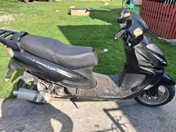 Czarny skuter z widocznym napisem TIGRA zaparkowany na betonowej powierzchni. W tle zielona trawa, drewniany pojemnik, czerwone ogrodzenie i cień skutera. Skuter posiada czarne siedzenie, wydech oraz kierownicę z lusterkami.