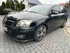 Czarny samochód osobowy typu kombi zaparkowany na brukowanej nawierzchni. Pojazd znajduje się przed domem z białą elewacją i ciemnym dachem. W tle widoczne są zielone krzewy i drzewa. Samochód ma metalowe felgi, a na przodzie widoczna jest nieczytelna tablica rejestracyjna. Nawierzchnia składa się z szarych, kwadratowych kamieni.