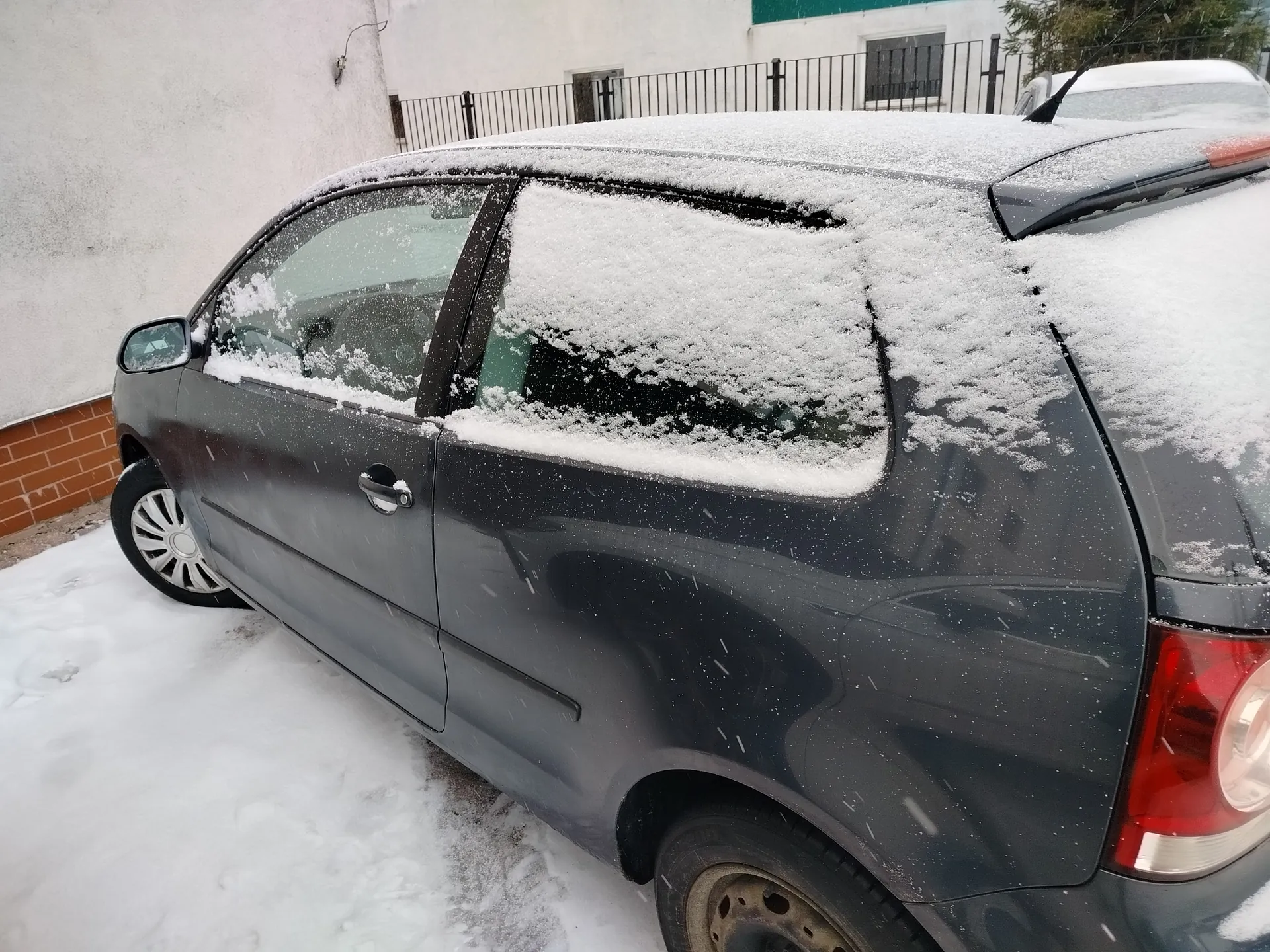 Ciemnoszary samochód osobowy pokryty warstwą śniegu, zaparkowany na śnieżnej powierzchni. W tle biały budynek z ceglaną podstawą, metalowe ogrodzenie oraz częściowo widoczny inny samochód. Pada śnieg.