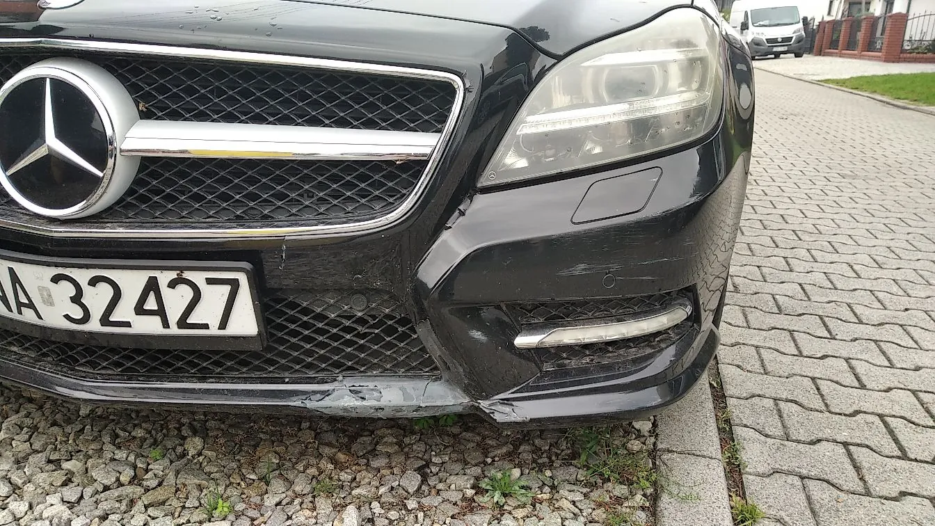 Zbliżenie na przód czarnego samochodu marki Mercedes-Benz. Widoczny jest charakterystyczny grill z logo marki oraz lewy reflektor. Zderzak po stronie kierowcy jest wyraźnie uszkodzony, z widocznymi wgnieceniami i zarysowaniami w dolnej części. Samochód posiada tablicę rejestracyjną z numerem A 32427. Pojazd stoi na krawężniku obok kostki brukowej.
