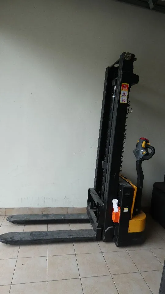Czarny wózek widłowy elektryczny z żółtymi elementami obudowy, stojący na jasnej, kafelkowej podłodze na tle białej ściany. Urządzenie posiada dwa długie, poziome widelce oraz pionowy maszt z czerwono-białymi znakami ostrzegawczymi. Po prawej stronie znajduje się rączka sterująca z czerwonym przyciskiem.