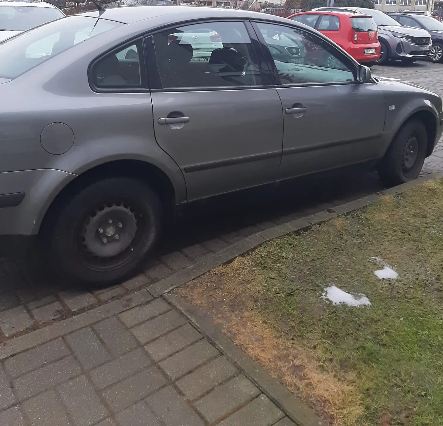 Szary samochód osobowy typu sedan zaparkowany na brukowanej powierzchni. Po prawej stronie widoczna trawa z resztkami śniegu. W tle inne pojazdy: czerwony samochód, srebrny i biały. Samochód na pierwszym planie ma czarne felgi i ciemne szyby. Tło obejmuje budynki i drzewa.