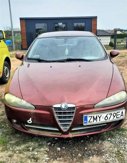 Czerwony samochód marki Alfa Romeo z przodu, z widoczną tablicą rejestracyjną ZMY 679EA. Po lewej stronie częściowo widoczny żółty samochód. Tło: budynek z niebiesko-brązowymi panelami, oknami i ogrodzeniem. Podłoga z drobnych kamieni i trawy. Niebo pochmurne.