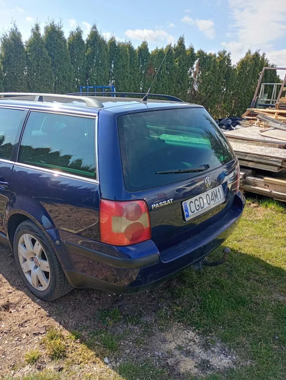 Tylne widok ciemnoniebieskiego samochodu osobowego Volkswagen Passat kombi z tablicą rejestracyjną CGD-04M1 (PL). Samochód stoi na podwórku z trawą i ziemią, w tle wysokie zielone drzewa, stos desek i niebieska konstrukcja. Widoczne są dachówka, reflektory oraz napis PASSAT na bagażniku.