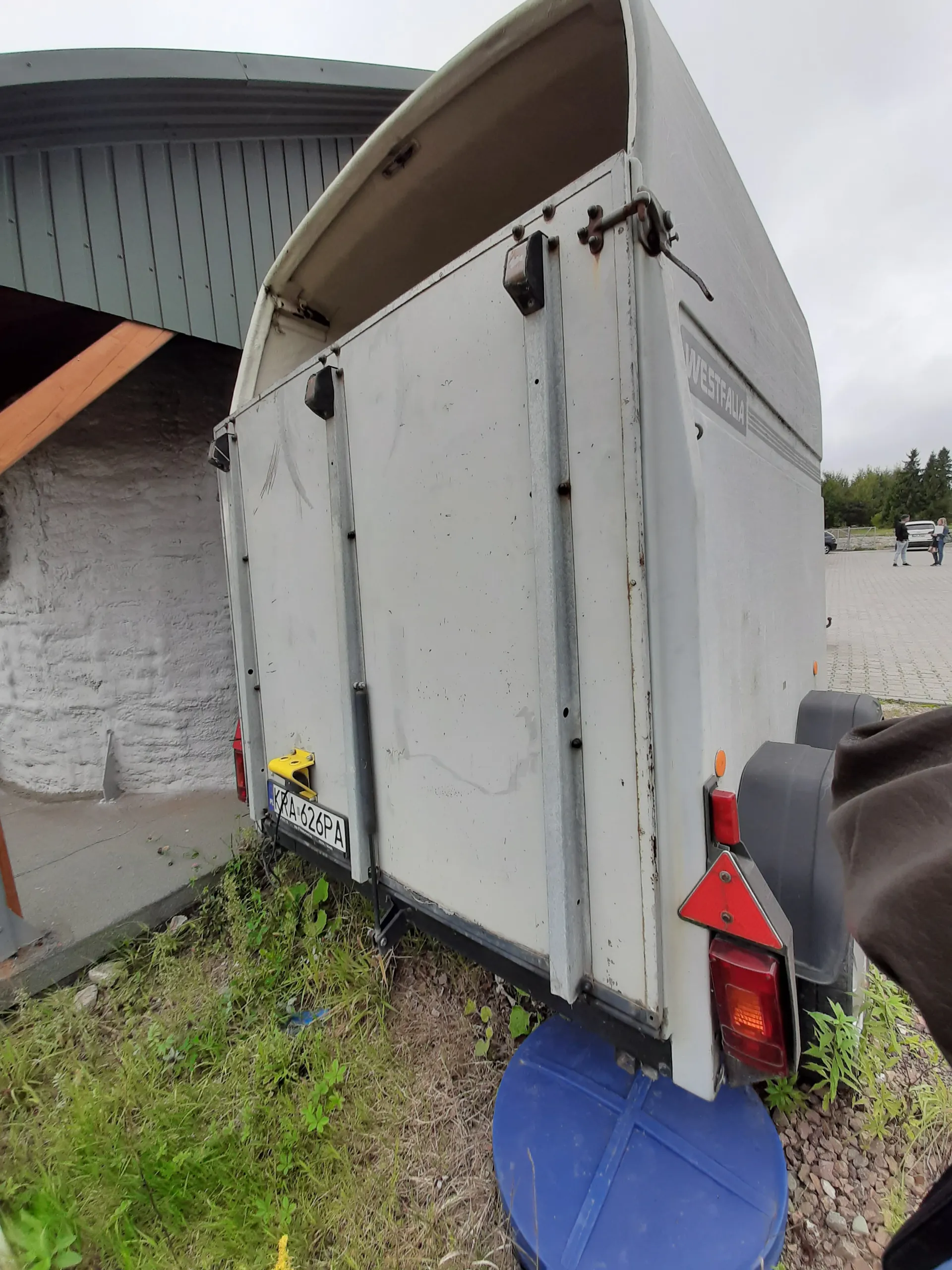 Zdjęcie przedstawia szarą przyczepę z napisem 'WESTFALIA' na boku. Widoczna jest tablica rejestracyjna 'KRA 628PA', czerwone światła tylnego oświetlenia oraz czerwony trójkątny odblask. Przyczepa stoi na trawie obok niebieskiego pojemnika. W tle znajduje się budynek z szarą blachą, chodnik, drzewa oraz osoba stojąca w oddali. Niebo jest zachmurzone.