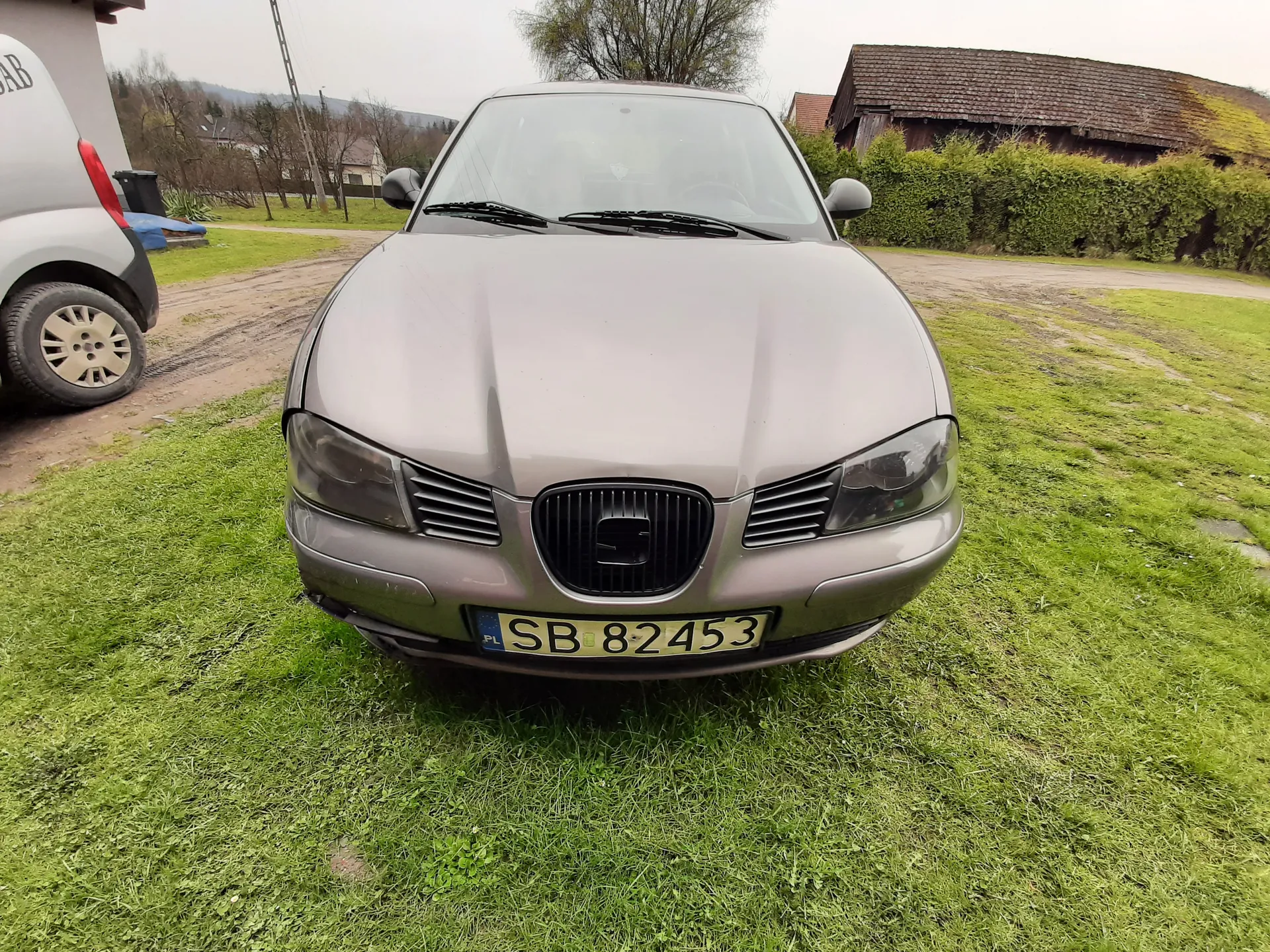 Przedni widok szarego samochodu marki Saab z tablicą rejestracyjną SB 82453 PL, stojącego na trawie. Po lewej stronie częściowo widoczny biały samochód z fragmentem napisu AB na tylnej szybie. W tle zielony żywopłot, domy, drzewa i niebo. Na tablicy rejestracyjnej widoczny napis SB 82453 oraz oznaczenie PL.