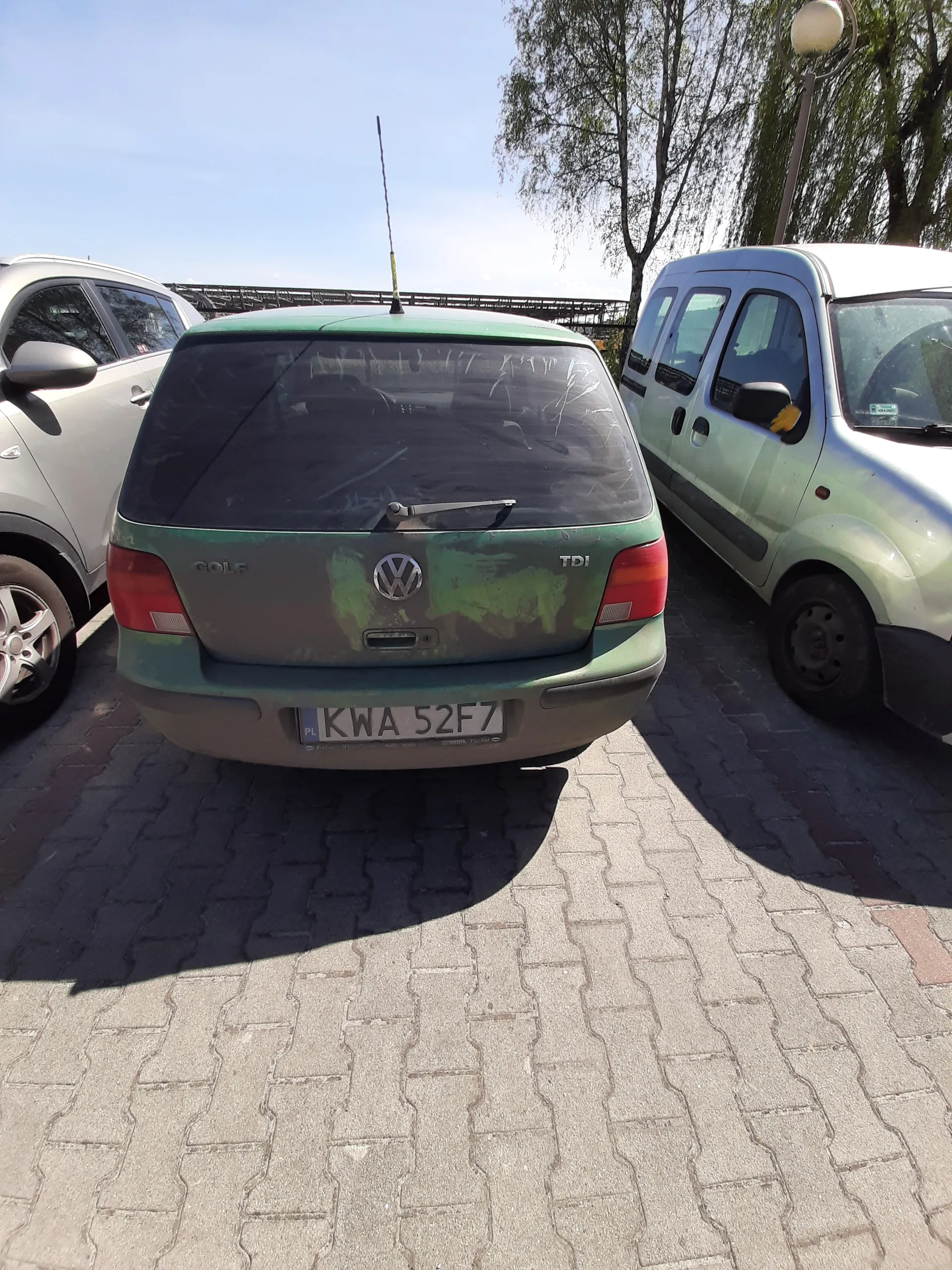 Trzy samochody zaparkowane na brukowanej powierzchni. W centrum zielony samochód Volkswagen Golf z tablicą rejestracyjną KWA 52F7, napisem GOLF i TDI na tylnej części. Obok niego biały samochód dostawczy oraz częściowo widoczny srebrny samochód osobowy. W tle drzewa, latarnia uliczna i niebo.