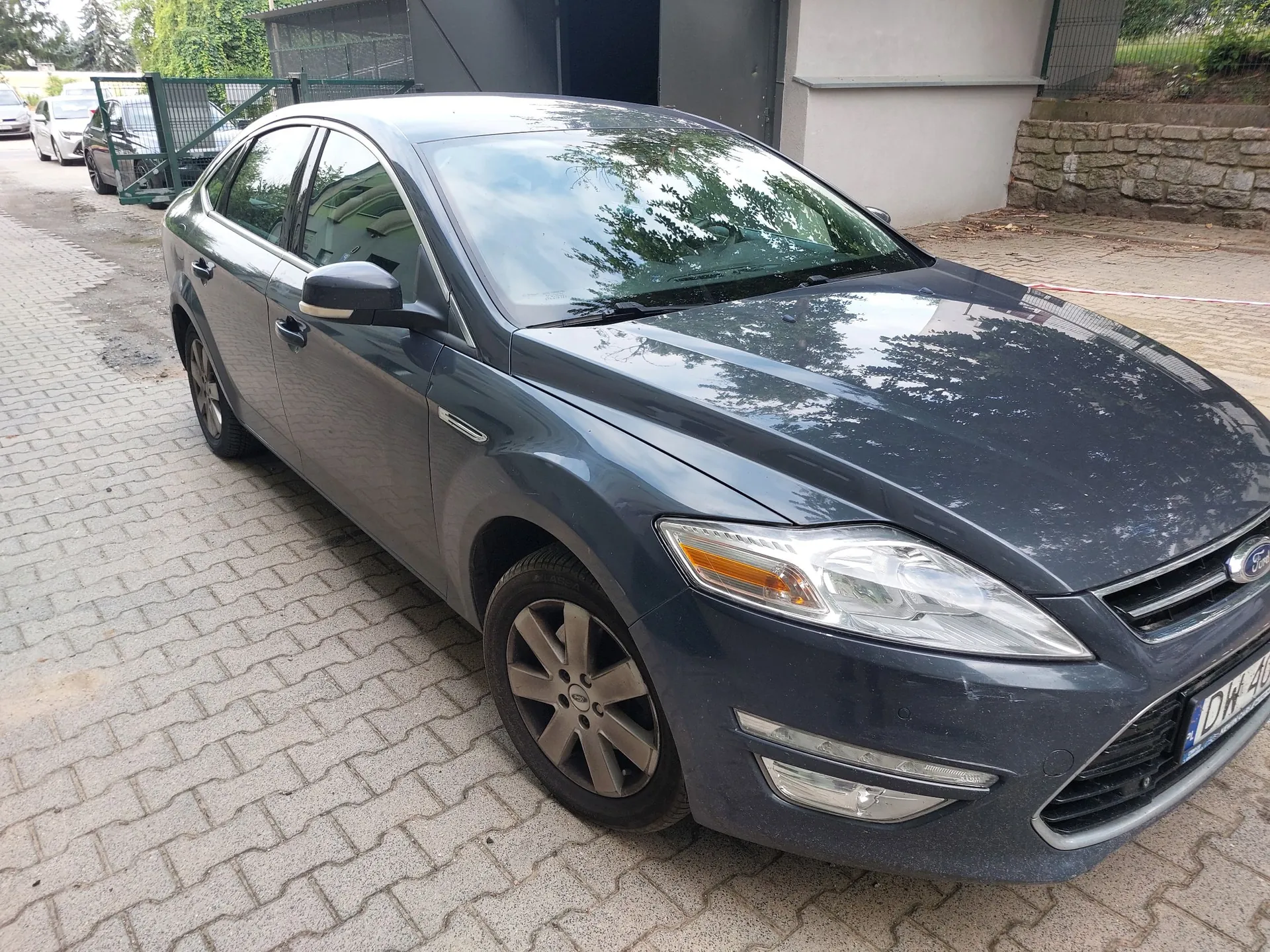 Ciemnoszary samochód osobowy marki Ford Mondeo zaparkowany na kostce brukowej. Pojazd jest ukazany z przodu pod kątem, widoczna jest prawa strona nadwozia, przednia lampa oraz felga. W tle widać inne samochody, ogrodzenie i fragment budynku.