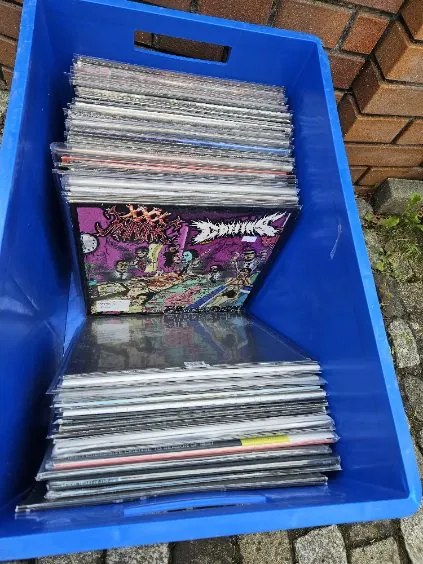 Niebieskie plastikowe pudełko wypełnione stosami winylowych płyt. Wśród nich widoczna okładka z kolorowym motywem i napisami 'Sodom' oraz 'Mortal Way of Live'. Tło stanowi ceglana ściana i brukowana posadzka.