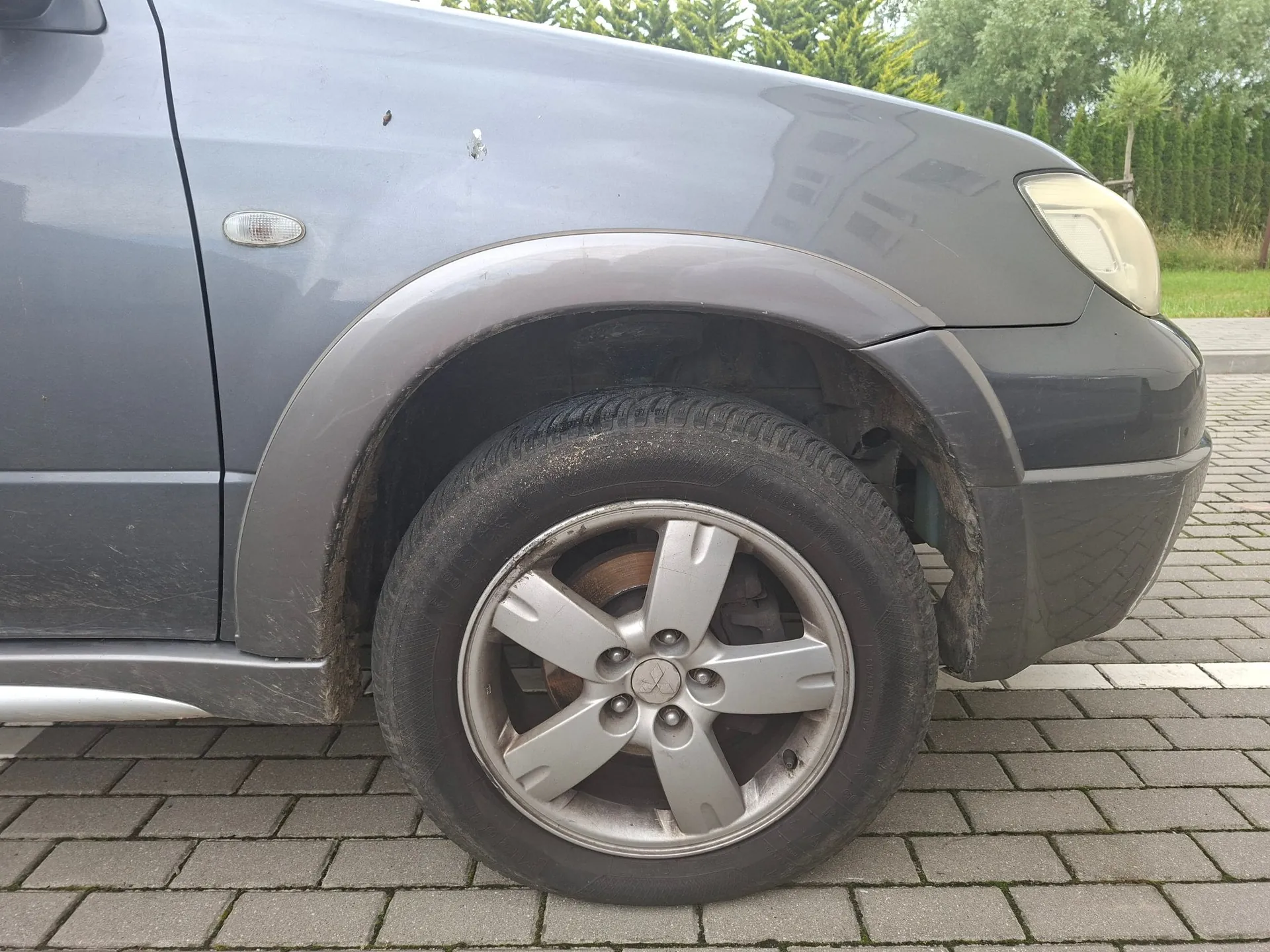 Zbliżenie na przednie prawe koło szarego samochodu zaparkowanego na kostce brukowej. Widoczny jest srebrny felga aluminiowa z pięcioma ramionami oraz fragment nadkola i przedniego reflektora. Na karoserii nad kołem widoczne są drobne zarysowania i zabrudzenia.