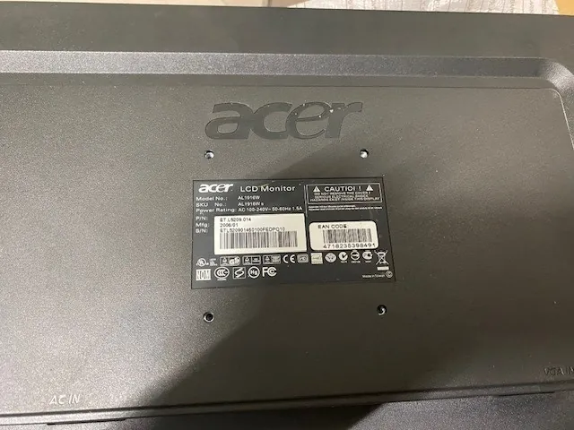 Tylna część czarnego monitora LCD marki Acer. W centralnej części znajduje się czarna etykieta z białym tekstem, w tym 'Acer LCD Monitor', 'K222HQL', 'CAUTION', 'ACER INC.' oraz kodami kreskowymi. Na etykiecie widoczne są symbole certyfikacji, takie jak CE, FCC i inne. Po prawej stronie obrazu umieszczono duży napis 'acer'. W lewym górnym rogu widoczny jest napis 'AC IN'.