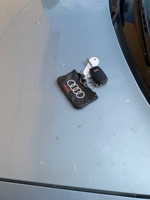 Kluczyki do samochodu z czarnym pilotem i białym brelokiem, leżące obok czarnego, skórzanego etui z logo Audi i napisem 'Audi' w kolorze czerwonym. Przedmioty znajdują się na jasnoszarej, lekko zakurzonej powierzchni maski samochodu.