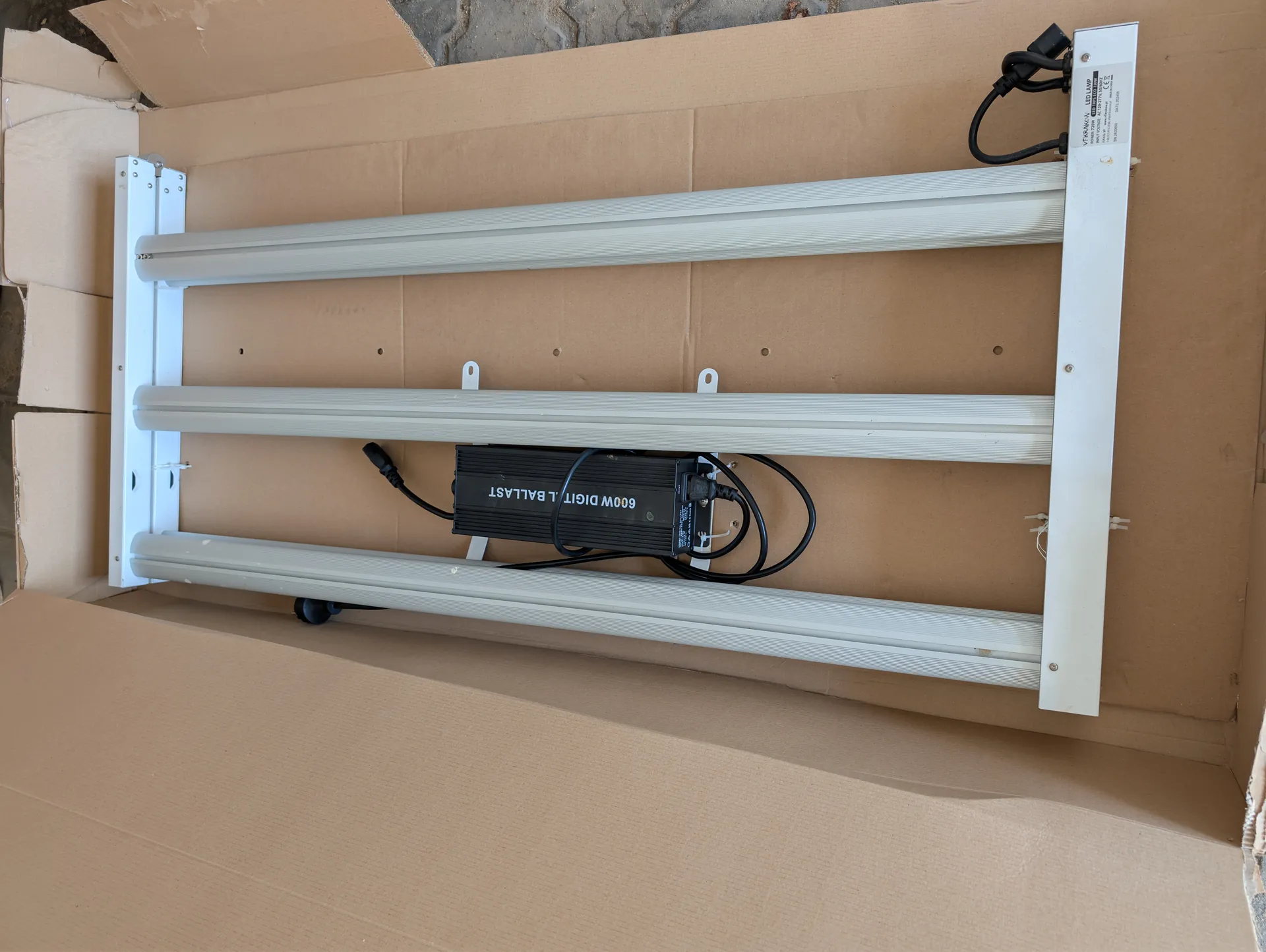 Otwarte kartonowe pudełko zawierające trzy długie, cylindryczne lampy LED zamocowane na białej ramie. W środku znajduje się czarny zasilacz z napisem '600W DIGITAL BALLAST' oraz przewody elektryczne. Na dolnej części ramy widoczna jest biała etykieta z dodatkowymi informacjami.