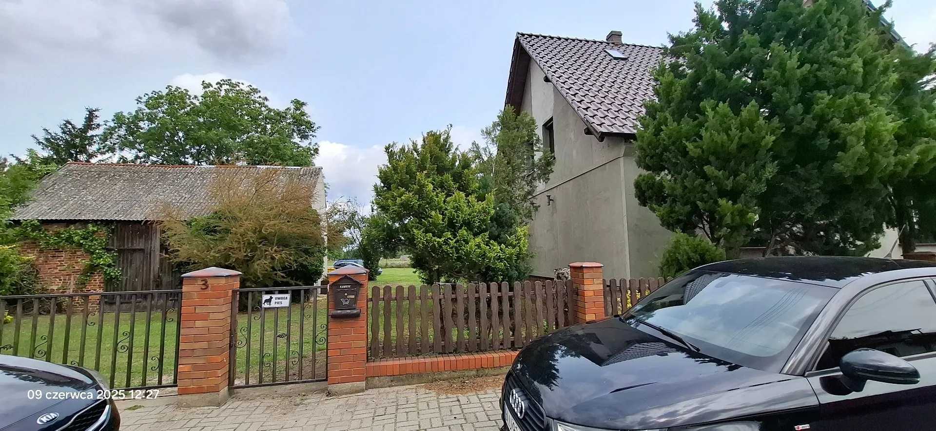Zdjęcie przedstawiające posesję z dwoma budynkami. Po lewej stronie znajduje się niski budynek gospodarczy z ceglanymi ścianami i dwuspadowym dachem pokrytym szarą dachówką. Po prawej stronie stoi wyższy, tynkowany dom z brązowym, spadzistym dachem. Posesja jest ogrodzona brązowym drewnianym płotem z ceglanymi słupkami. Na jednym ze słupków widnieje numer 3, a na bramie tabliczka z napisem 'UWAGA PIES'. W prawym dolnym rogu widoczny jest fragment czarnego samochodu marki Audi. W tle widać zielone drzewa i krzewy pod zachmurzonym niebem. W lewym dolnym rogu znajduje się data '09 czerwca 2025 12:27'.