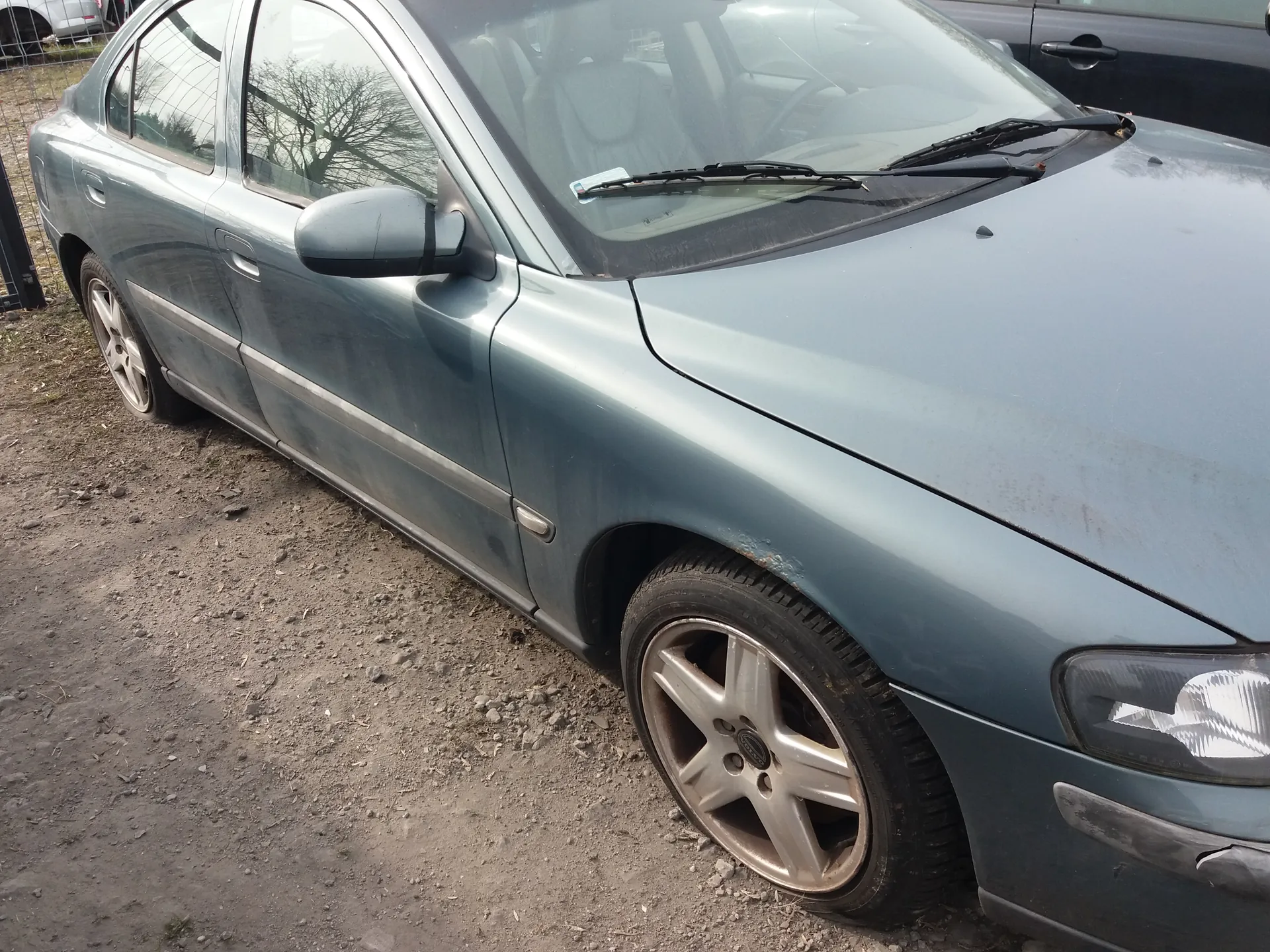 Niebieski samochód osobowy typu sedan zaparkowany na podłożu z drobnej ziemi i kamieni. Pojazd ma widoczne zabrudzenia na karoserii, zwłaszcza w dolnej części. Widoczne są przednie koło z aluminiową felgą, reflektor, lusterko boczne oraz przednie wycieraczki. W tle fragment ogrodzenia, drzewa bez liści oraz część innego pojazdu. Na przedniej szybie znajduje się mały niebieski znaczek.