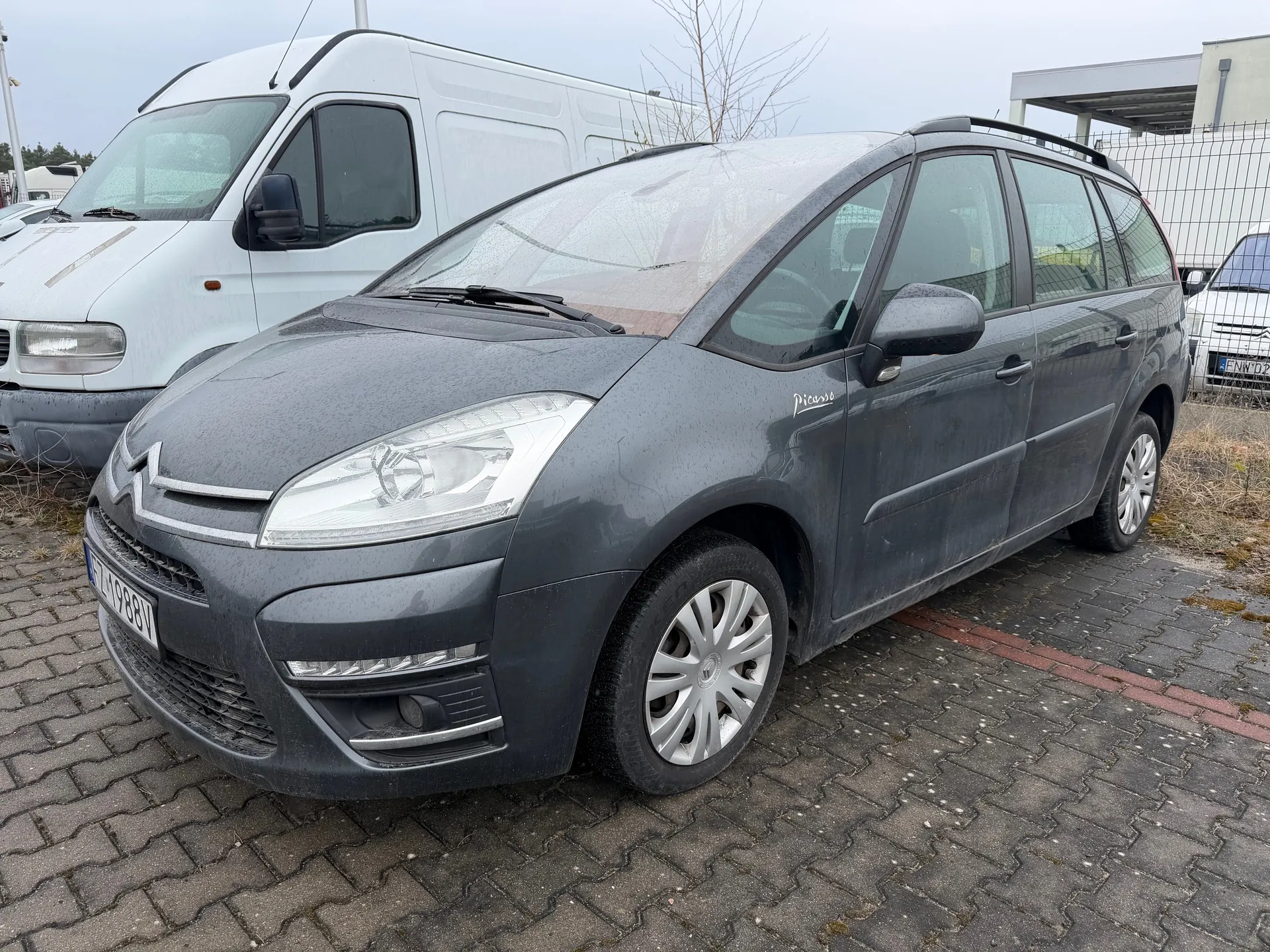 Szary samochód Citroën Picasso z tablicą rejestracyjną ZL 988V zaparkowany na brukowanej powierzchni. W tle biały van, inne pojazdy, ogrodzenie i budynki. Niebo jest zachmurzone. Na boku samochodu widoczny napis 'Picasso'.