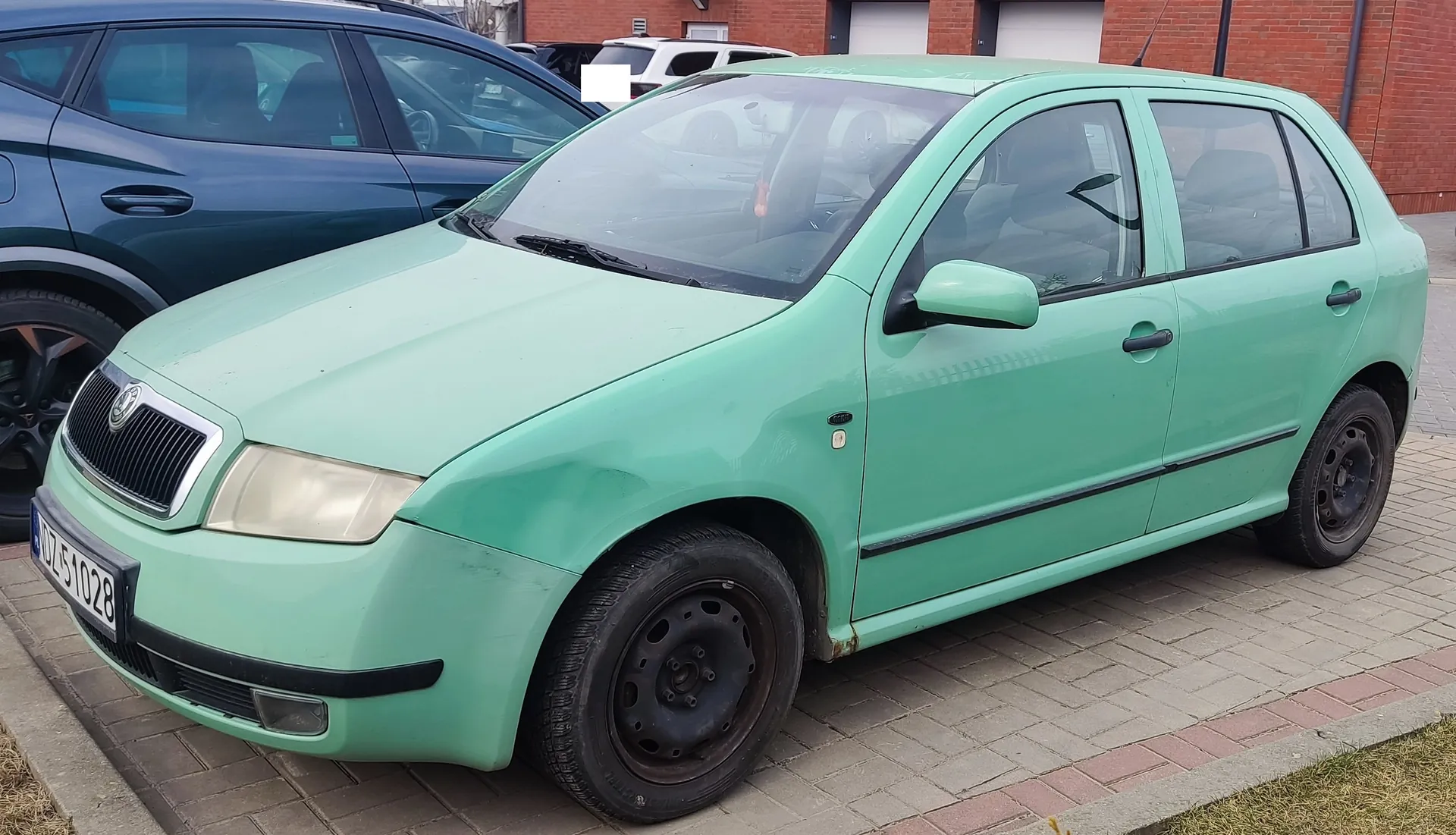 Zielony samochód osobowy marki Škoda, model Fabia, zaparkowany na brukowanej powierzchni. Widoczna tablica rejestracyjna DZ51028. Samochód ma czarne felgi, lekko zabrudzoną karoserię i uszkodzony przedni zderzak. W tle ciemnoniebieski samochód oraz ceglany budynek z białymi bramami garażowymi. Na pierwszym planie fragment trawnika.
