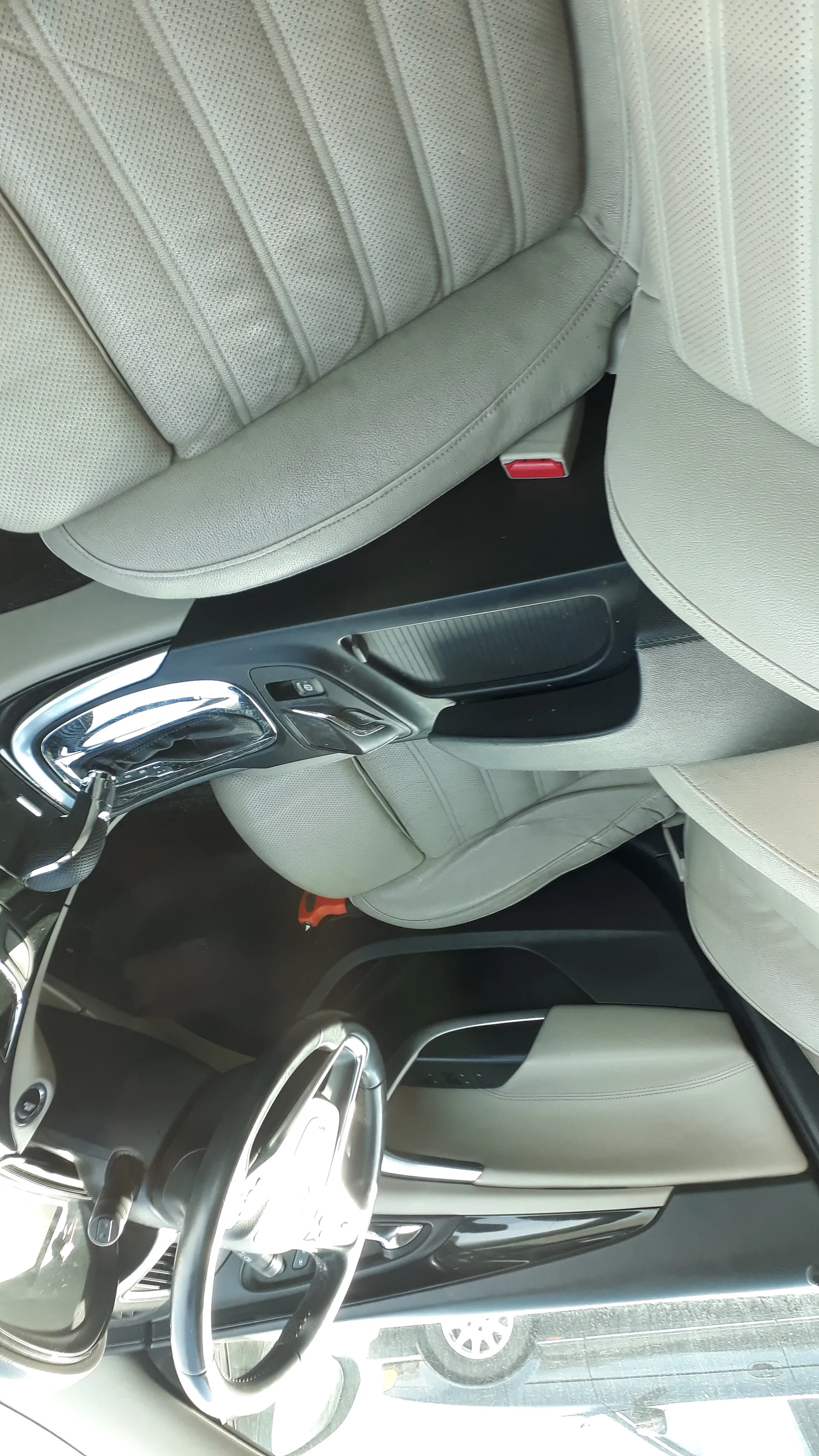 Wnętrze samochodu z jasnoszarymi skórzonymi fotelami o perforowanej powierzchni. Widoczna kierownica po lewej stronie, centralna konsola z dźwignią zmiany biegów oraz fragment lusterka bocznego. Po lewej stronie obrazu widoczny fragment zewnętrzny z częścią innego pojazdu. Brak czytelnego tekstu.