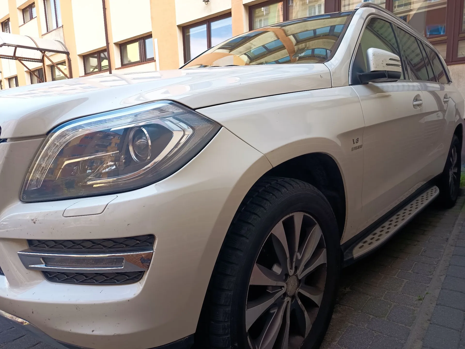 Zbliżenie na przód i prawy bok białego samochodu typu SUV marki Mercedes-Benz. Widoczny jest reflektor, przednie prawe koło z wieloramienną felgą oraz stopień boczny. Na błotniku znajduje się emblemat 'V6 BLUEEFFICIENCY'. W tle widoczny jest fragment wielopiętrowego budynku mieszkalnego z oknami.