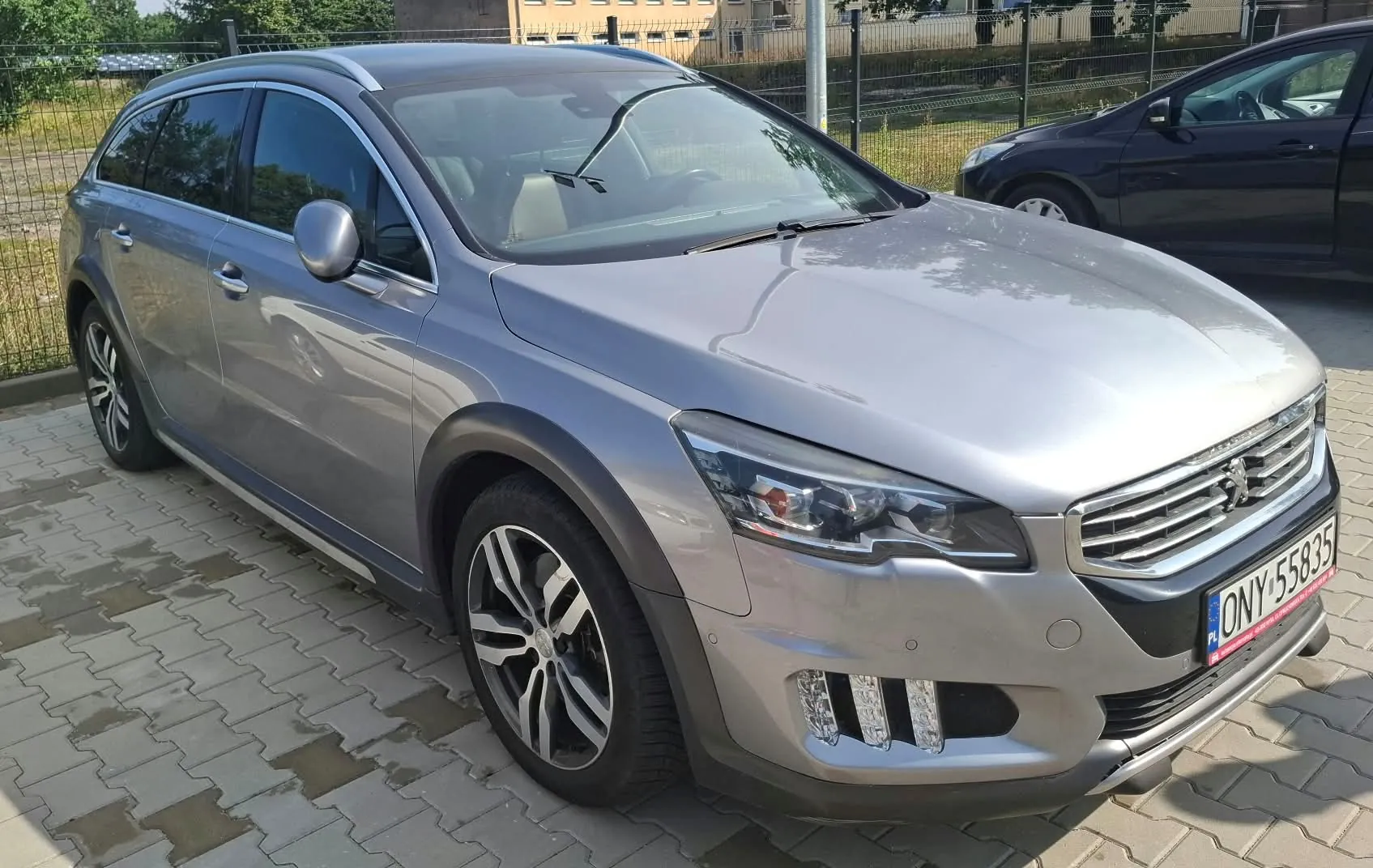 Srebrny samochód osobowy typu kombi marki Peugeot zaparkowany na kostce brukowej. Pojazd jest widoczny z przodu i z boku. Na samochodzie widoczna jest polska tablica rejestracyjna o numerze ONY 55835. W tle widać czarny samochód oraz ogrodzenie z siatki.