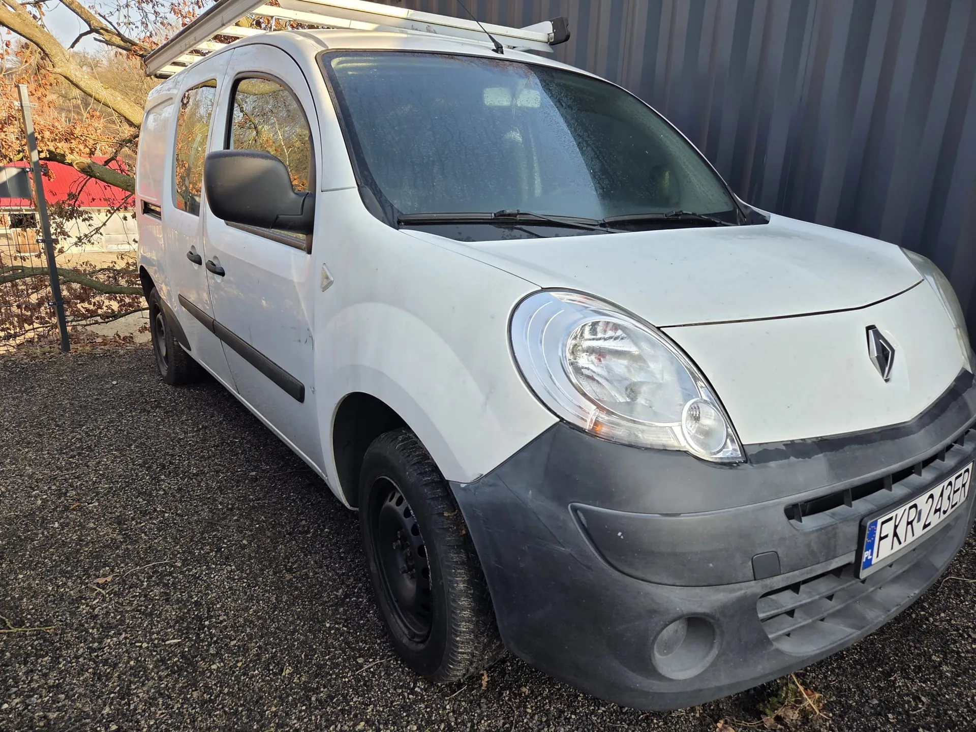 Biały samochód dostawczy marki Renault z tablicą rejestracyjną FKR 243ER (PL) zaparkowany na żwirowym podłożu. Pojazd posiada czarny zderzak, dachowy uchwyt na ładunek oraz lusterko boczne. W tle widoczne są drzewa z suchymi liśćmi, metalowa budowla oraz czerwony dach budynku.