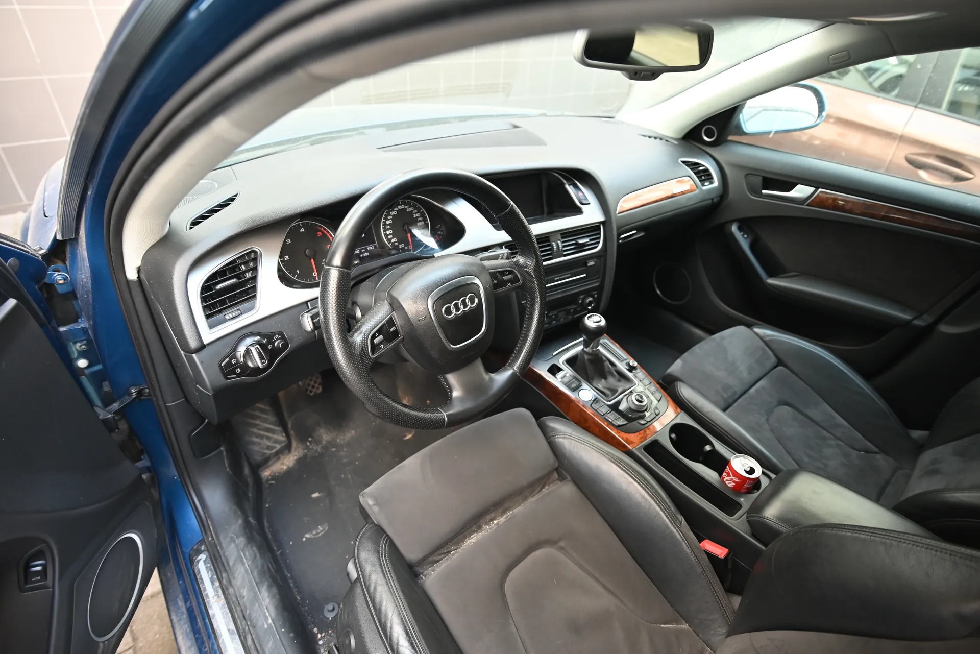 Wnętrze samochodu marki Audi z widocznym kierownicą z logo czterech pierścieni, tablicą przyrządów, czarnymi skórzonymi fotelami i centralną konsolą. Drzwi po stronie kierowcy są otwarte, a w uchwycie na napoje znajduje się puszka Coca-Cola. Wnętrze posiada drewniane wstawki na desce rozdzielczej i drzwiach. Podłoga wydaje się brudna. W tle widoczne są elementy budynku i inny pojazd.