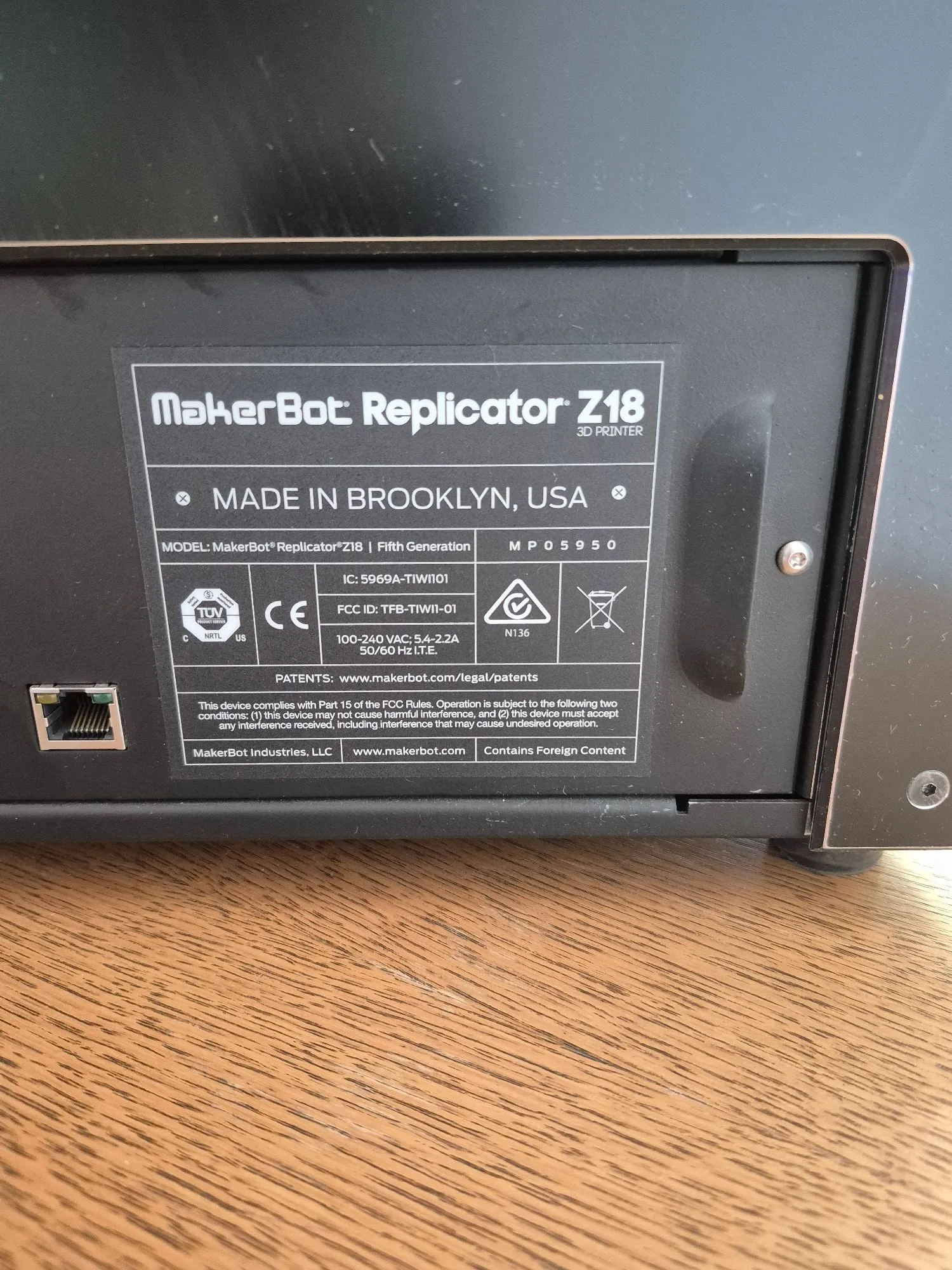 Zdjęcie tabliczki znamionowej drukarki 3D MakerBot Replicator Z18. Na czarnej etykiecie znajduje się biały tekst: 'MakerBot Replicator Z18 3D PRINTER', 'MADE IN BROOKLYN, USA', 'MODEL: MakerBot Replicator Z18 | Fifth Generation', numer seryjny 'MP05950', 'IC: 5969A-TIW1101', 'FCC ID: TFB-TIW11-01'. Widoczne są certyfikaty TOV, CE oraz oznaczenia elektryczne: '100-240 VAC; 5.4-2.2A 50/60 Hz I.T.E.'. Na dole znajduje się informacja o zgodności z FCC oraz adres strony internetowej www.makerbot.com i nazwa firmy MakerBot Industries, LLC.