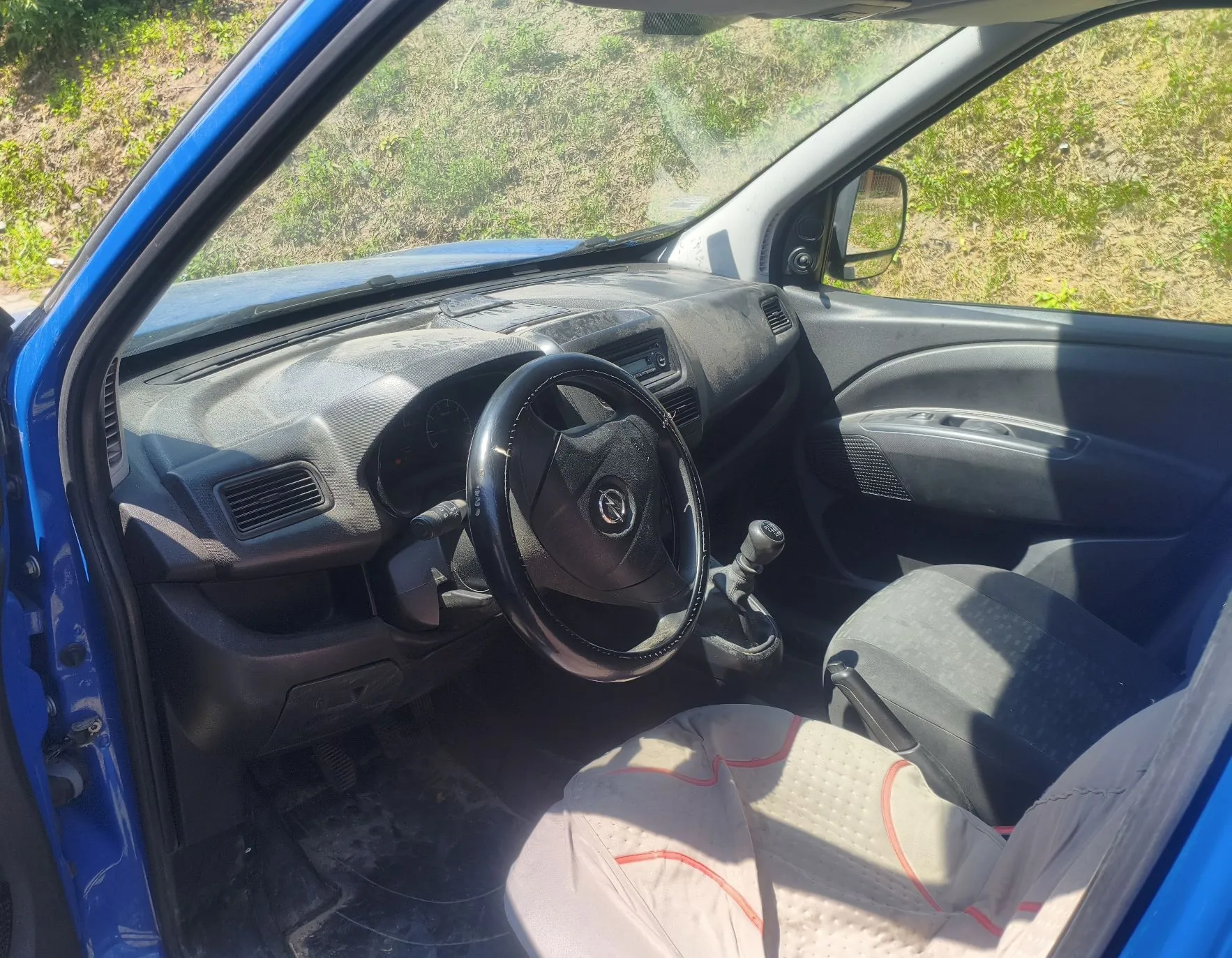 Wnętrze niebieskiego samochodu widziane z otwartych drzwi pasażera. Widoczna jest zakurzona deska rozdzielcza, czarna kierownica z logo marki Opel, lewarek zmiany biegów oraz dwa przednie fotele. Fotele są przykryte jasnymi nakładkami z czerwonymi przeszyciami. Przez przednią szybę widoczny jest zielony, trawiasty teren.