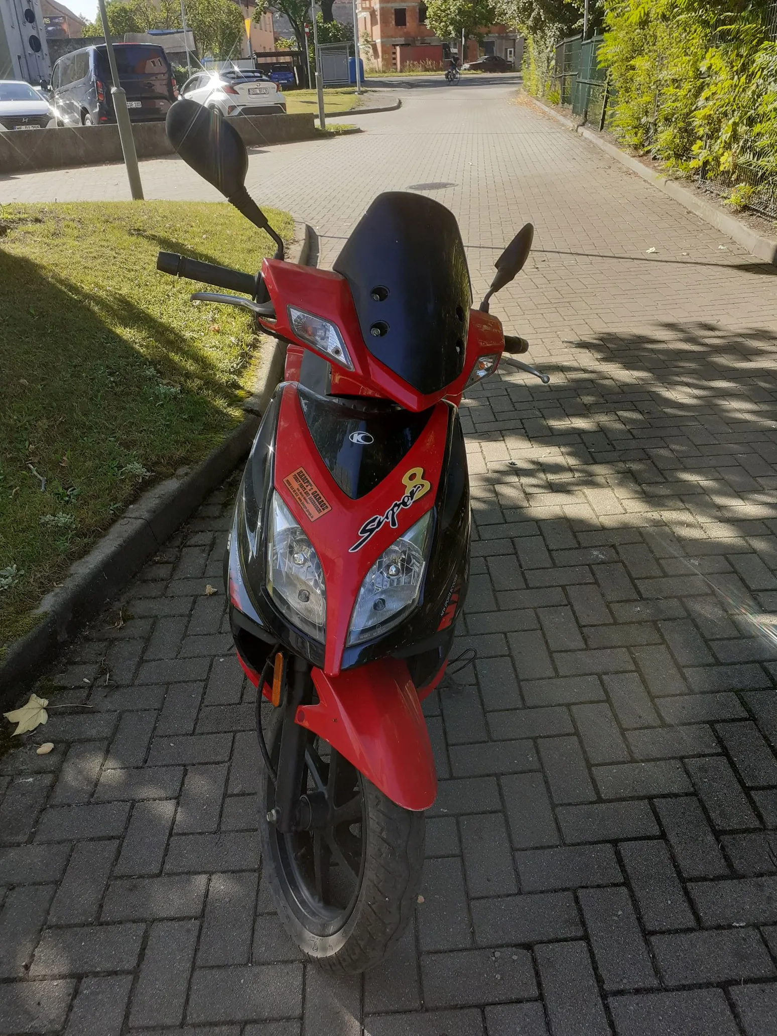 Czerwono-czarny skuter zaparkowany na kostce brukowej. Na przedniej owiewce widnieje napis 'S-max' oraz mniejsza naklejka 'HAPPY'S GARAGE'. Skuter jest ustawiony bokiem do obiektywu, w tle widoczny jest chodnik, trawnik oraz fragmenty budynków.