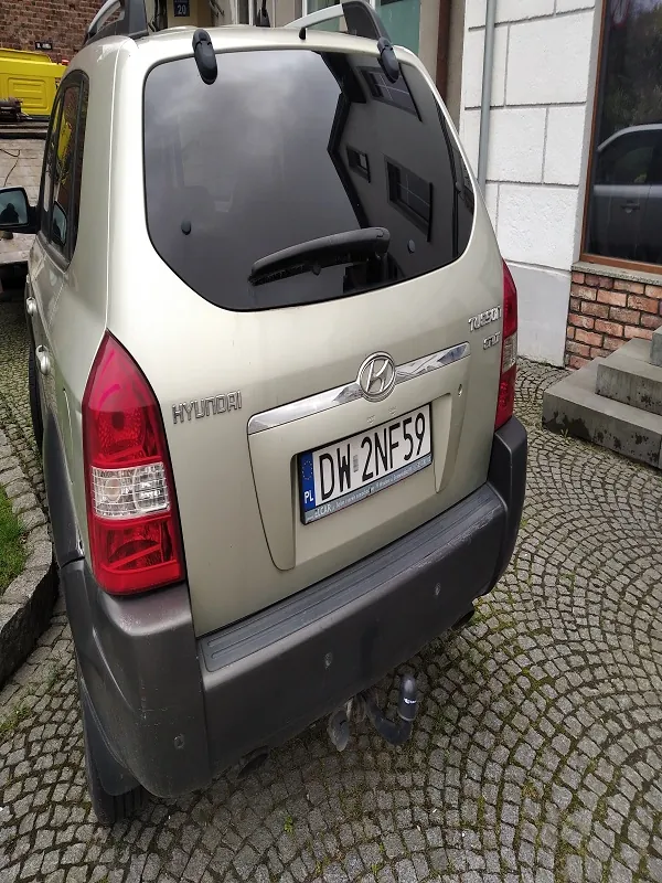 Z tyłu widoczny srebrny samochód marki Hyundai Tucson z tablicą rejestracyjną DW 2NF59 (PL). Samochód stoi na brukowanej powierzchni z okrągłym wzorem. W tle budynek z białymi płytkami i ceglaną częścią pod oknem, a także fragment żółtego pojazdu po lewej stronie. Na tylnym szybie widoczny wycieraczka, a na bagażniku logo marki i napis 'Tucson'.