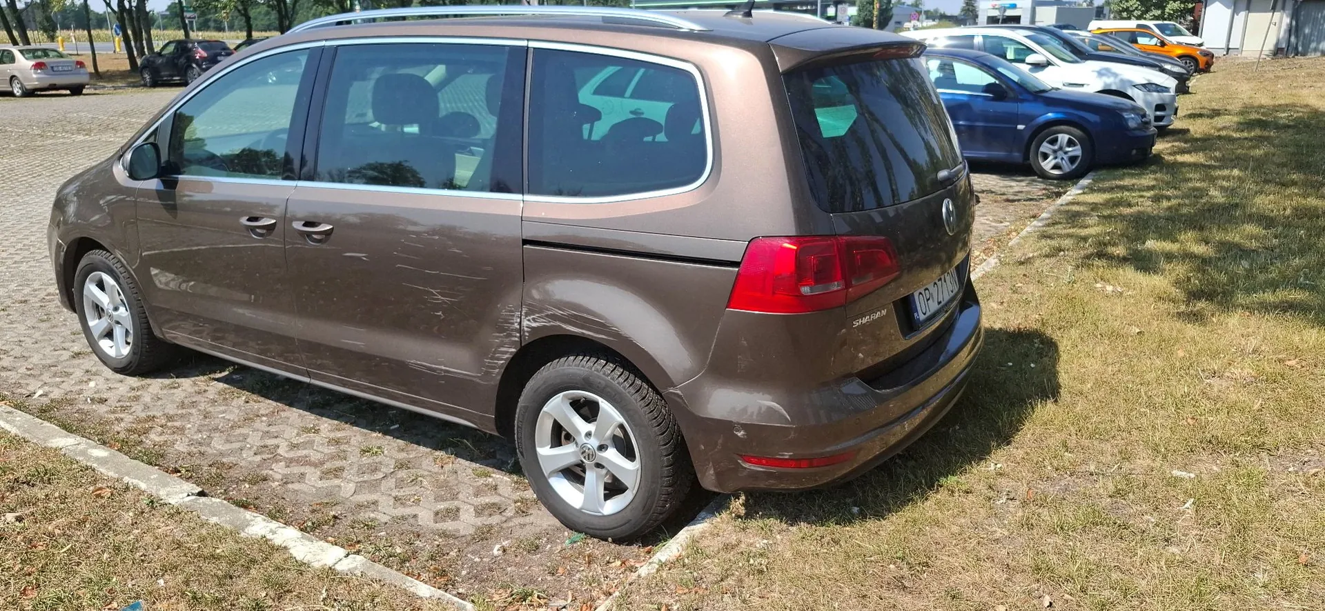 Brązowy samochód typu minivan marki Volkswagen Sharan zaparkowany na trawiastym placu. Pojazd posiada widoczne zarysowania i wgniecenia na prawym boku, na drzwiach oraz błotniku. W tle widoczne są inne zaparkowane samochody w kolorach niebieskim, białym i pomarańczowym oraz fragmenty infrastruktury drogowej.