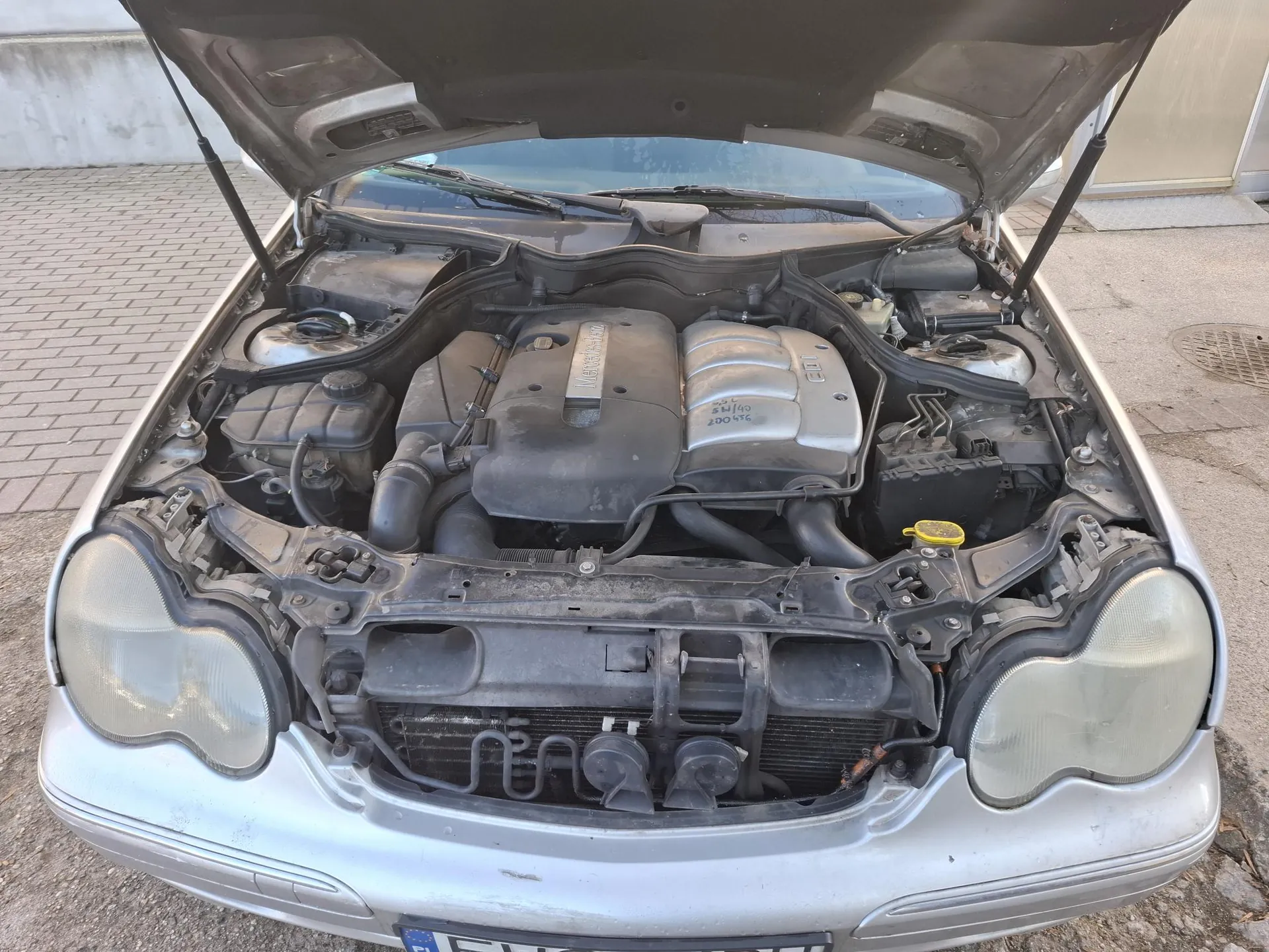 Srebrny samochód z otwartą maską, ukazujący silnik. W silniku widoczne są elementy takie jak pokrywa z napisem "Mercedes-Benz", oznaczenie "CDI" oraz cyfry "200 456". Samochód stoi na brukowanej powierzchni, w tle budynek i klapa kanalizacyjna. Na dole widoczna część tablicy rejestracyjnej z nieczytelnym tekstem.