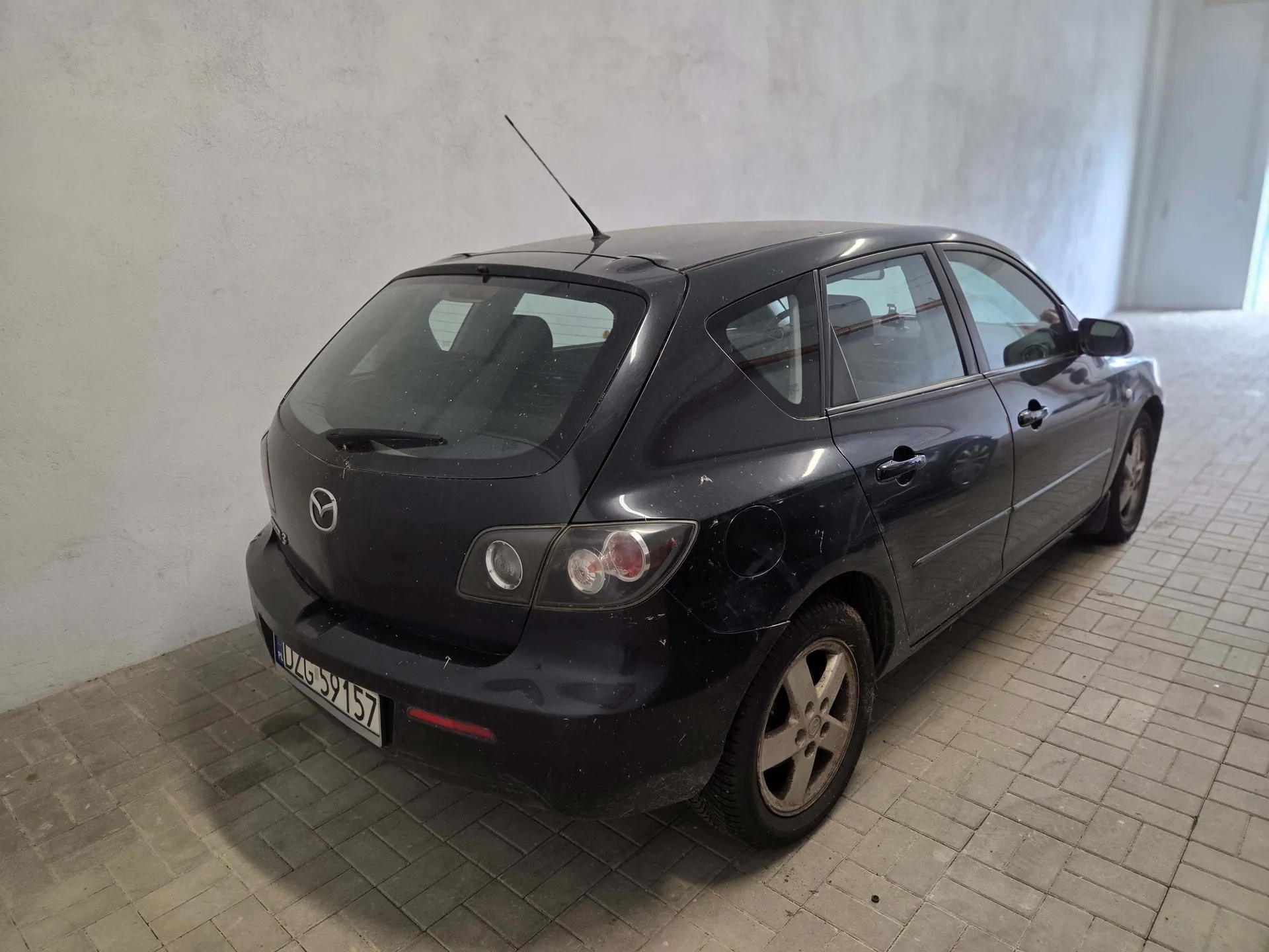 Czarny samochód marki Mazda w nadwoziu hatchback, zaparkowany w jasnym pomieszczeniu o białych ścianach i posadzce z kostki brukowej. Pojazd jest widoczny od tyłu i z boku, posiada tablicę rejestracyjną o numerze LU 04G 59157.