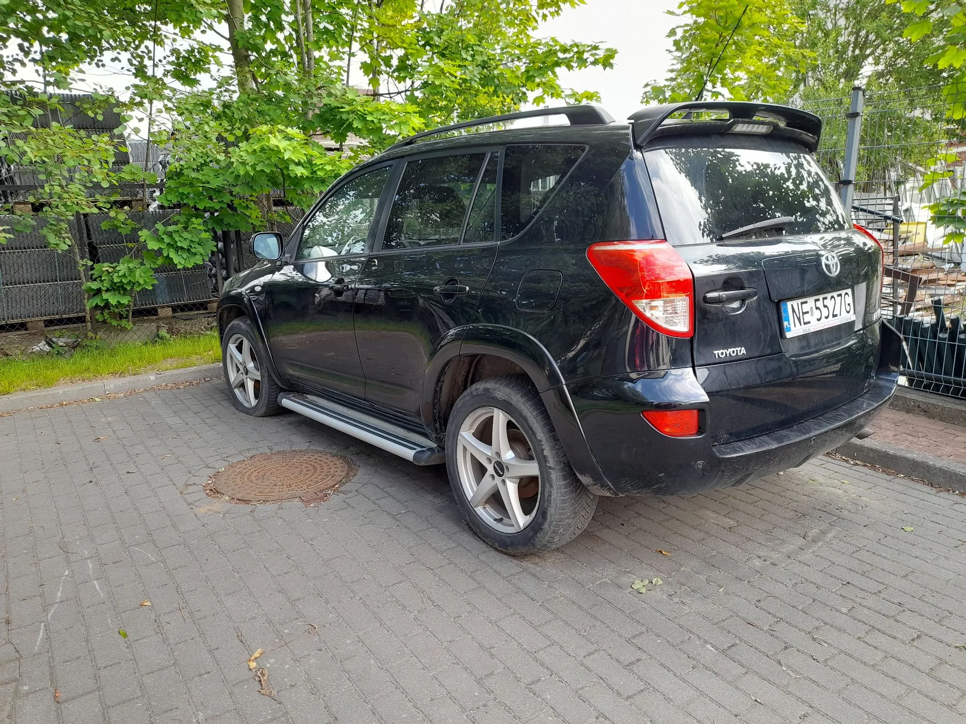 Czarny samochód marki Toyota typu SUV zaparkowany na kostce brukowej. Pojazd jest widoczny z tyłu i z boku. Na klapie bagażnika znajduje się napis TOYOTA oraz tablica rejestracyjna o numerze NE 5527G. W tle widoczne są zielone drzewa i fragmenty ogrodzenia.