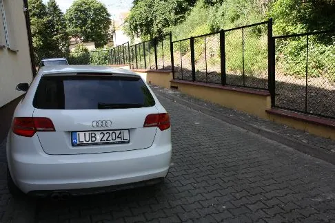 Tył białego samochodu marki Audi zaparkowanego na kostce brukowej. Samochód ma tablicę rejestracyjną o numerze LUB 2204L. W tle widoczny jest czarny metalowy płot z siatką, osadzony na niskim żółtym murku, oraz zielone drzewa i krzewy.