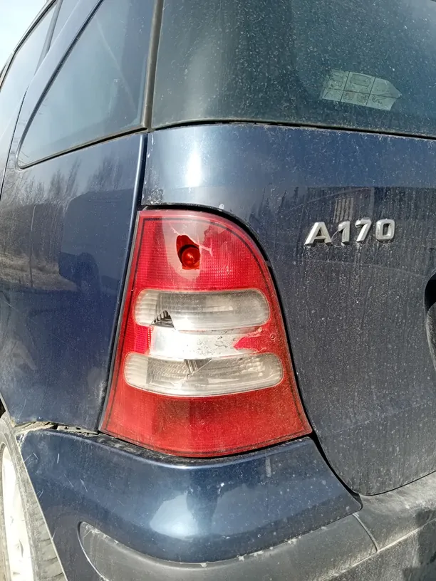 Zbliżenie na tylną część ciemnoniebieskiego samochodu. Widoczny czerwony reflektor z trzema poziomymi sekcjami oraz napis 'A170' na bagażniku. Samochód jest zabrudzony kurzem, a w tle odbijają się drzewa i budynki.