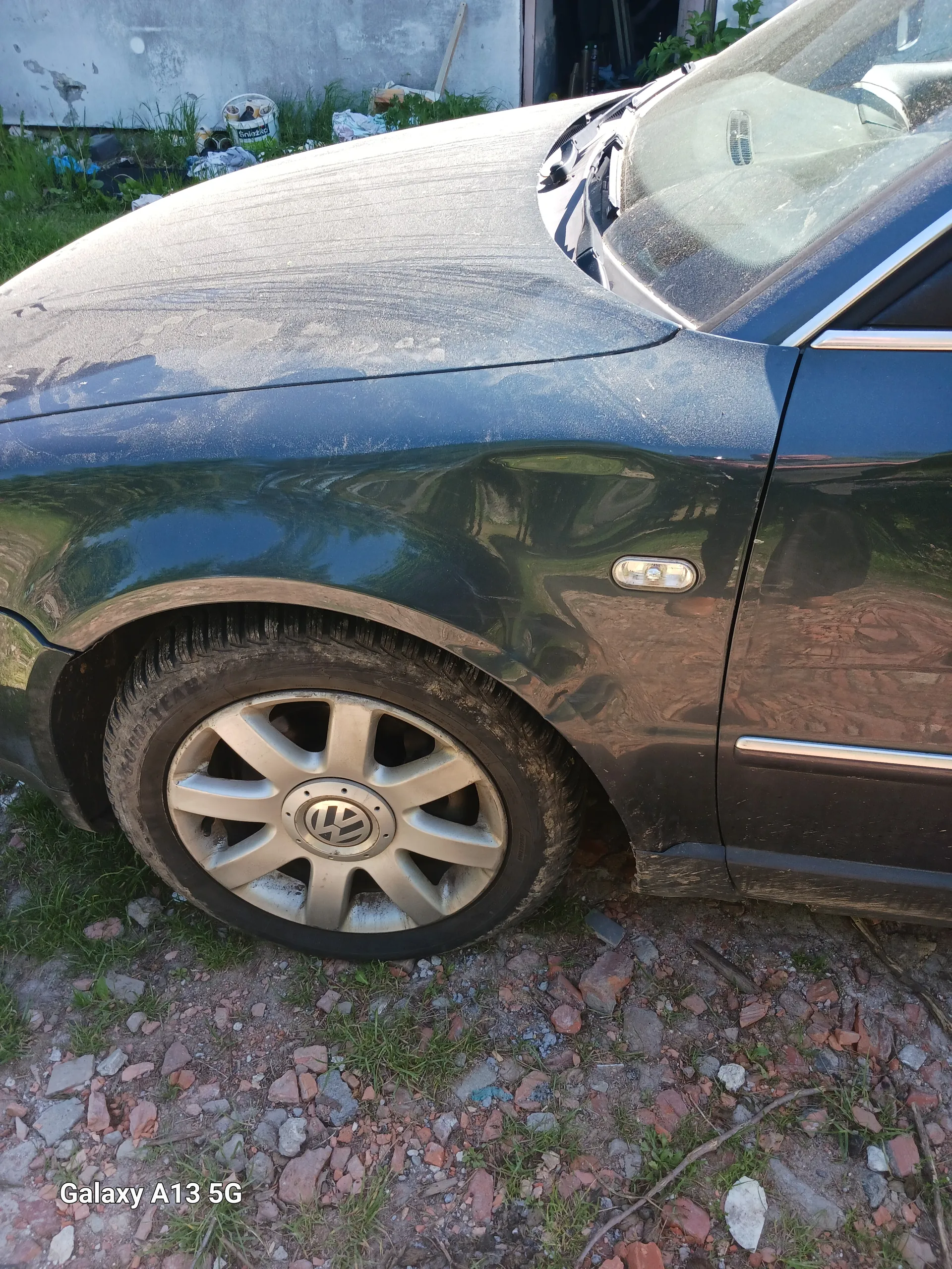 Czarny samochód marki Volkswagen, widok przedniej części z kołem z logo VW. Widoczne reflektory, brud na karoserii oraz podłoga z kamieniami i trawą. Po prawej stronie obrazu widoczny pionowy tekst 'Galaxy A13 5G'.