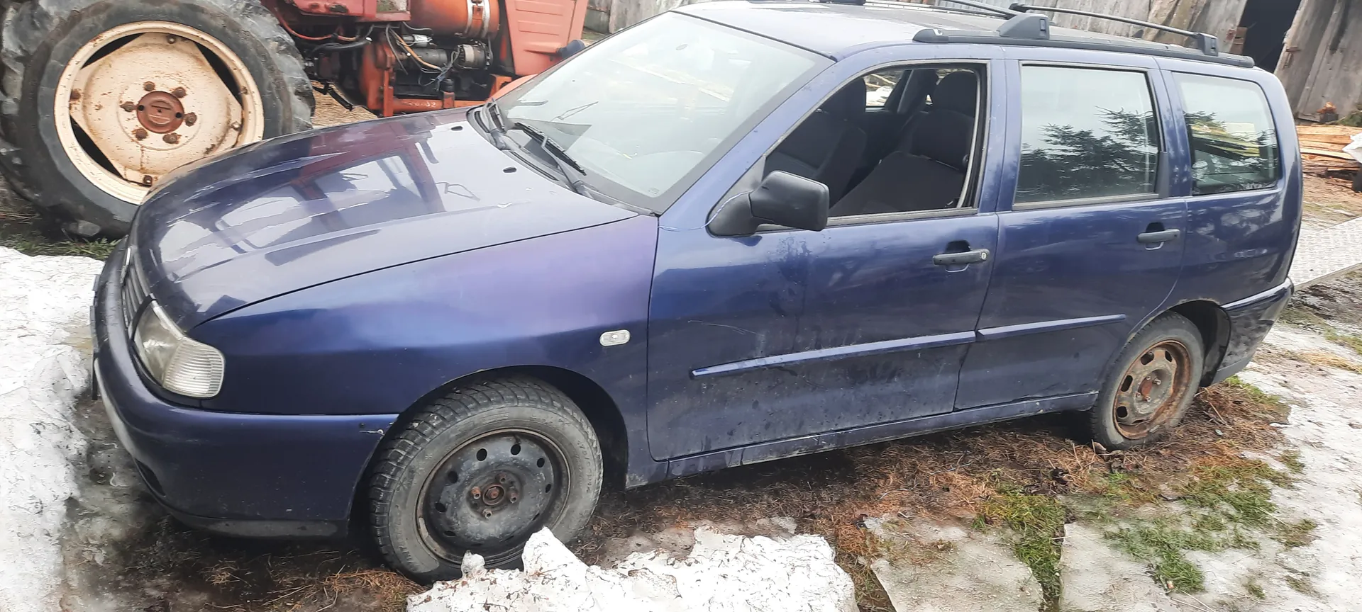 Niebieski samochód kombi z brakującym przednim kołem, zardzewiałymi felgami i zabrudzoną karoserią. W tle czerwony traktor z dużym kołem, na ziemi resztki śniegu i trawa. Samochód stoi na nierównym podłożu z widocznymi śladami zużycia.