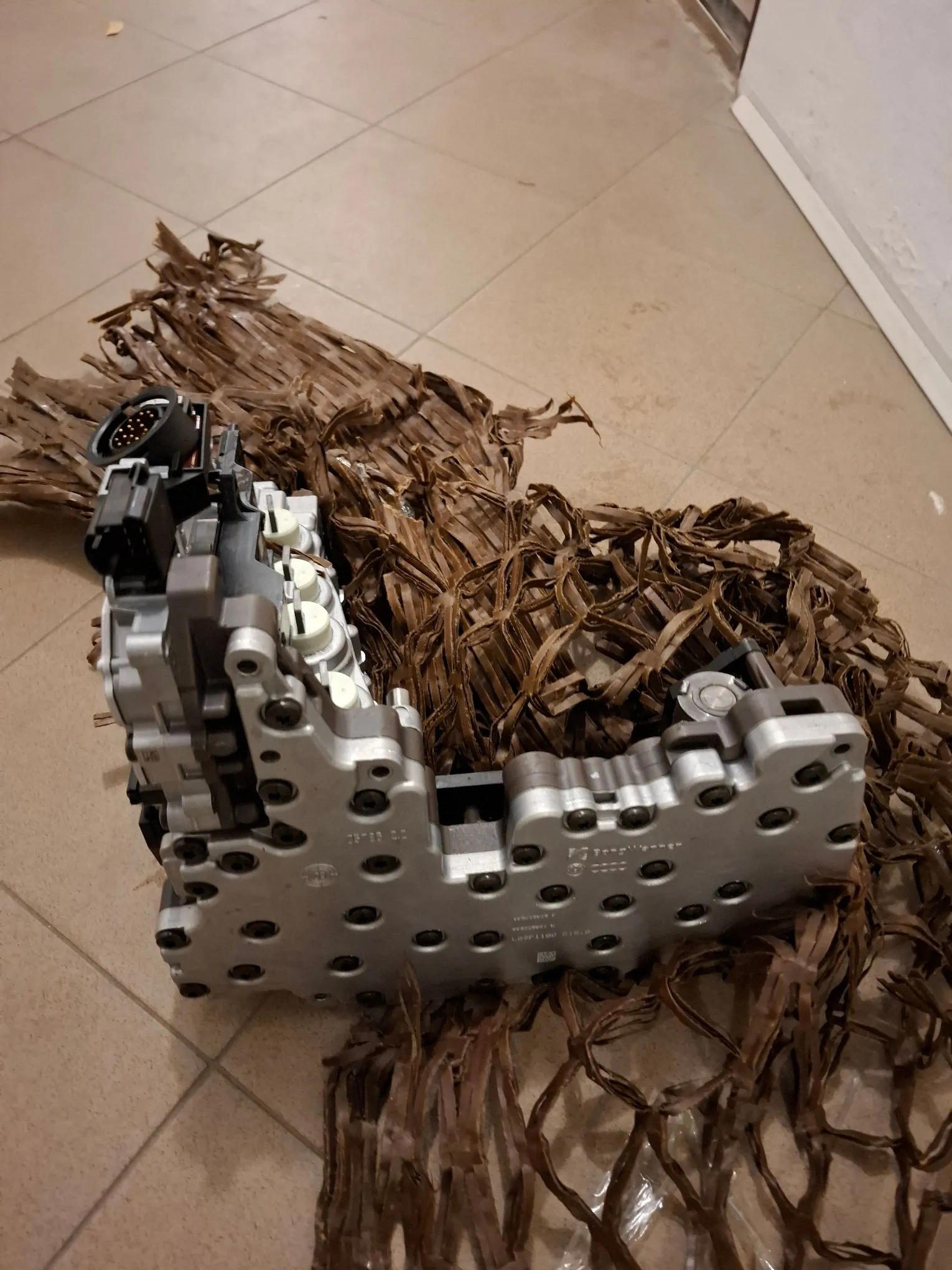 Metalowa część mechaniczna, prawdopodobnie moduł elektroniczny lub element zawieszenia samochodu, leżąca na brązowej, plecionej macie z włókien naturalnych na kafelkowej podłodze. Część jest srebrna, z widocznymi śrubami, złączem elektrycznym z wieloma pinami oraz nadrukami technicznymi na obudowie.