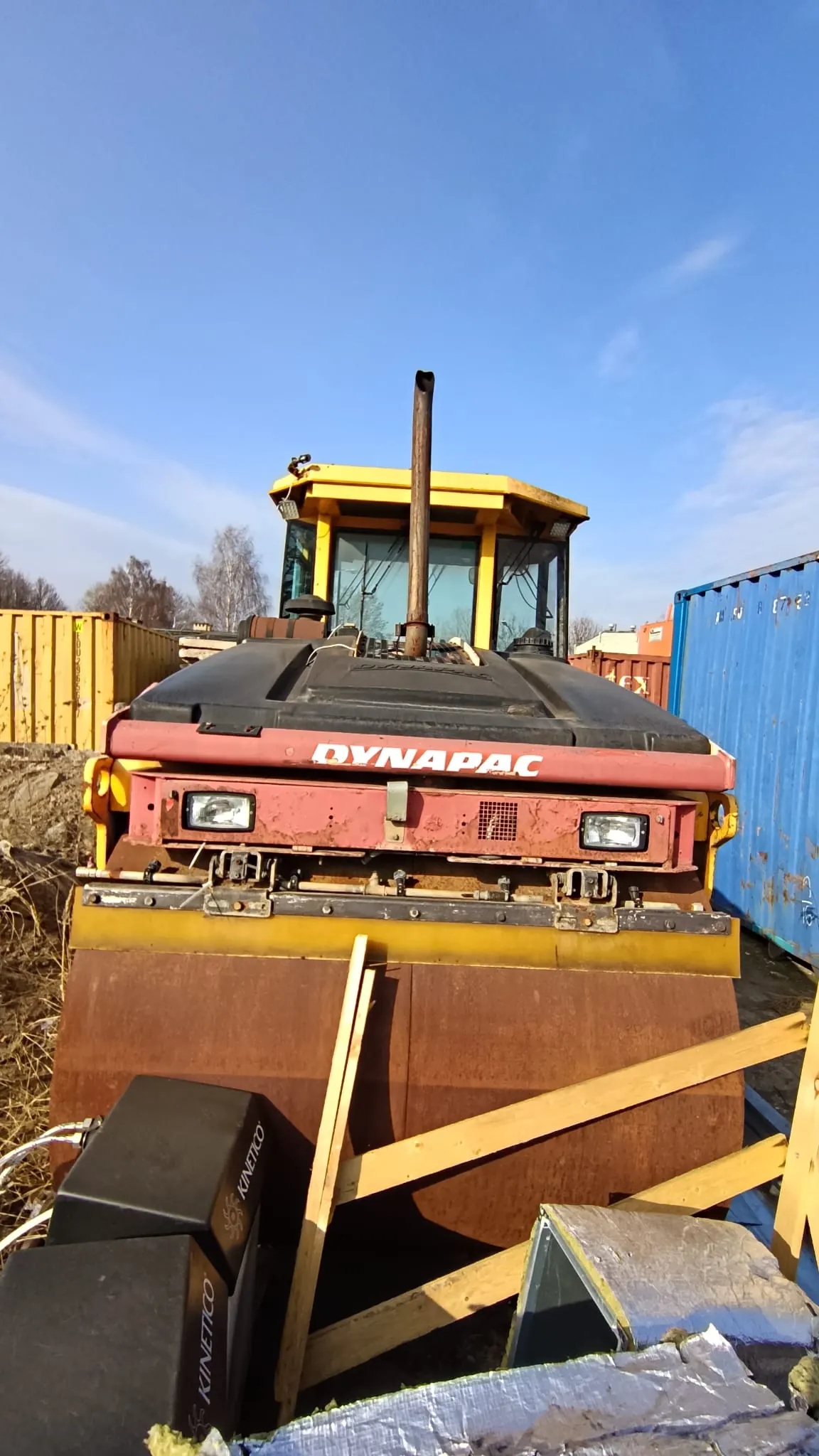 Zdjęcie przedstawia ciężką maszynę budowlaną, prawdopodobnie walec drogowy marki Dynapac. Maszyna ma żółtą kabinę operatora i czerwoną obudowę silnika z białym napisem „DYNAPAC”. Z przodu znajduje się szeroki, metalowy, rdzawy wałek zagęszczający, który jest częściowo osłonięty drewnianymi deskami. Po prawej stronie widoczny jest niebieski kontener transportowy, a po lewej żółty. W tle widać bezchmurne, niebieskie niebo.